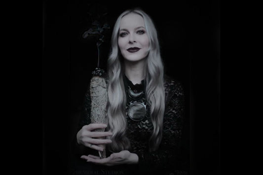 A black and white image of a blonde, witchy looking woman, Kari Dern of Become Spellbound is wearing a large mirror-like obsidian crystal necklace and holding a smoking sage bundle.