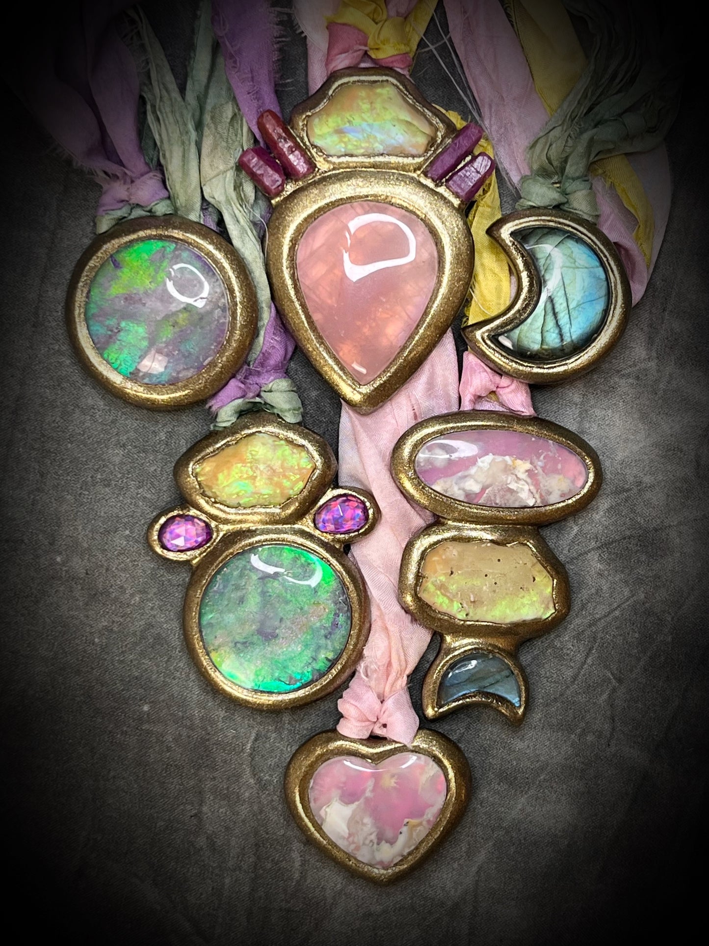 A handmade multi-piece necklace featuring various colorful opal and moonstone cabochons set in gold-toned metal, draped over pink and purple fabric against a dark textured background.