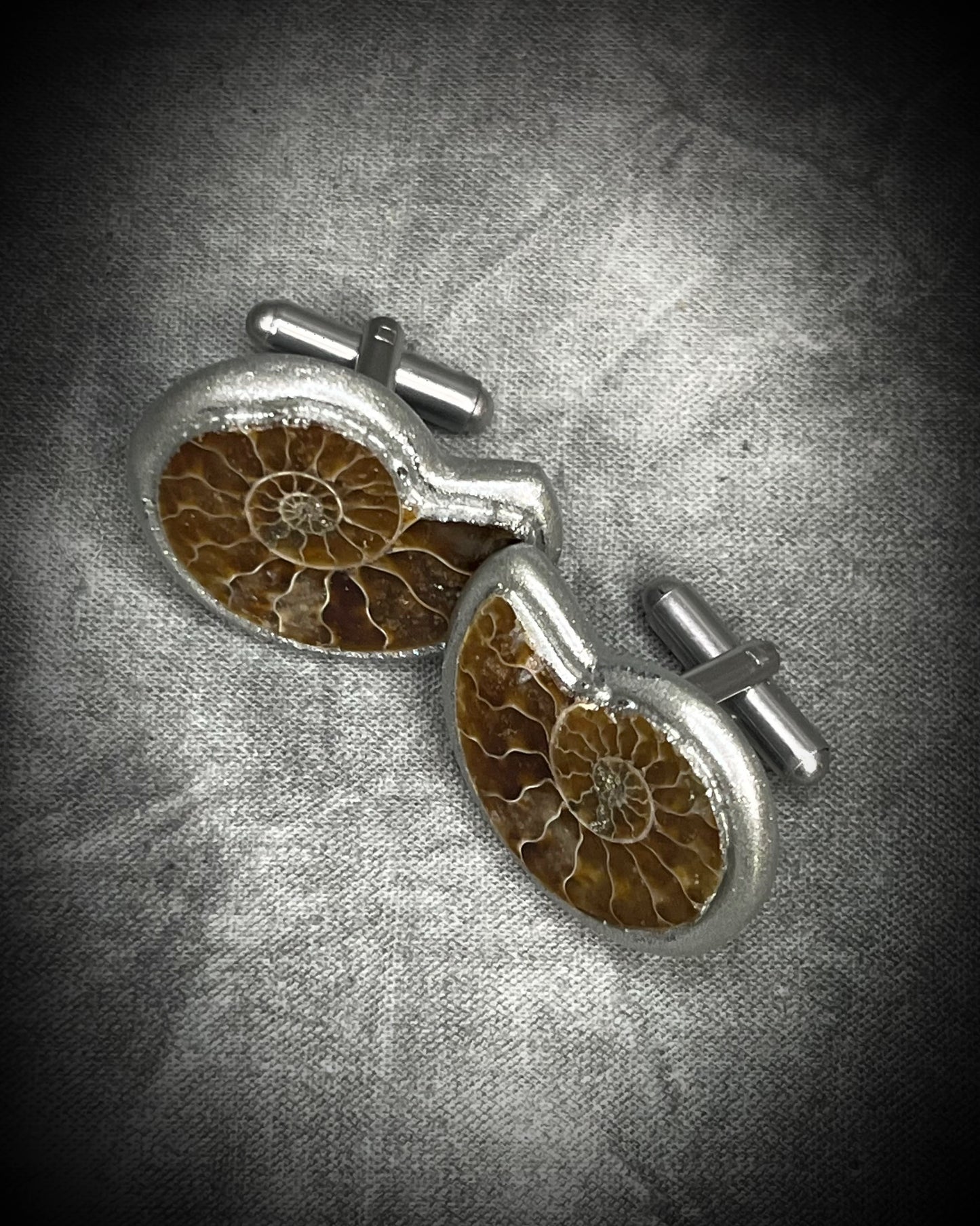 A pair of silver cufflinks featuring detailed brown and amber fossil inlays, displayed on a textured gray background.
