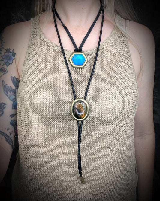 A person wearing a black cord bolo-style necklace with two pendants: a bright turquoise blue hexagonal pendant near the collarbone and a larger oval pendant with a brown and gold eye design at the lower center, shown against a dark background.
