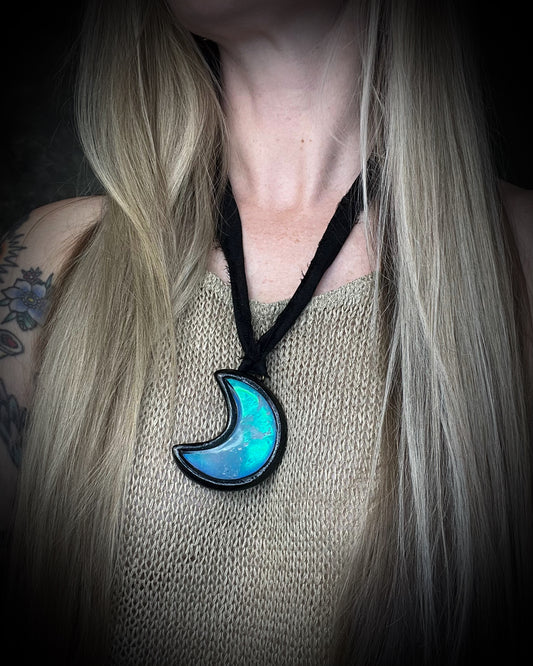 Quartz crescent moon crystal talisman with a luminous turquoise glow in a sculptural black-finish setting on black recycled silk, handmade by Become Spellbound in Baltimore, Maryland.
