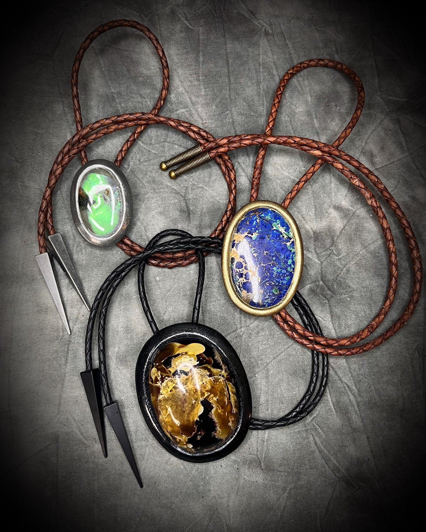 Three Bolo ties with blue, green and amber crystals and gunmetal silver and gold metal tips on brown and black braided leather. Boho western 2026 jewelry trend gift for her.