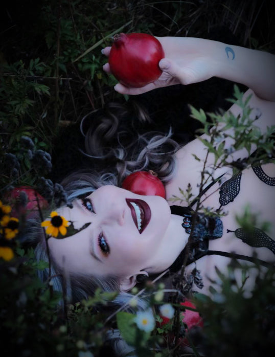 A fair woman with blonde hair, blue eyes and dark burgundy lipstick is wearing a black necklace and a bodysuit with snakes crawling up and laying in a field of yellow and white flowers, smiling and holding a pomegranate.