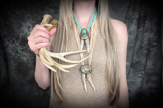  A woman with long blonde hair and a beige knit top is wearing layered necklaces featuring naturally shed deer antlers, glowing quartz gemstones and hung from recycled silk. She is also holding a few pairs of naturally shed antlers. 
