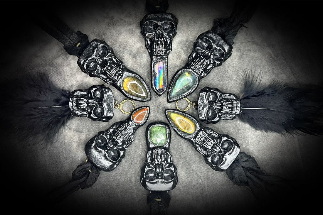 six black skull and gemstone necklaces and a pair of skull and feather earrings are arrayed in a circle with the gemstones pointing inward, hung alternately from leather or recycled silk