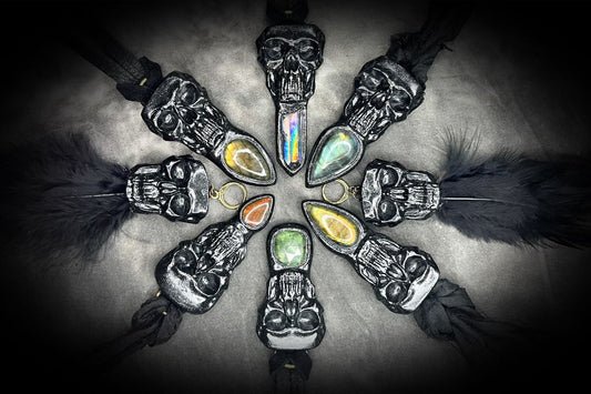 six black skull and gemstone necklaces and a pair of skull and feather earrings are arrayed in a circle with the gemstones pointing inward, hung alternately from leather or recycled silk