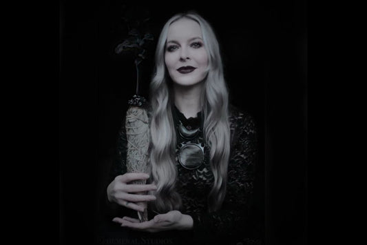 A black and white image of a blonde, witchy looking woman, Kari Dern of Become Spellbound is wearing a large mirror-like obsidian crystal necklace and holding a smoking sage bundle. 