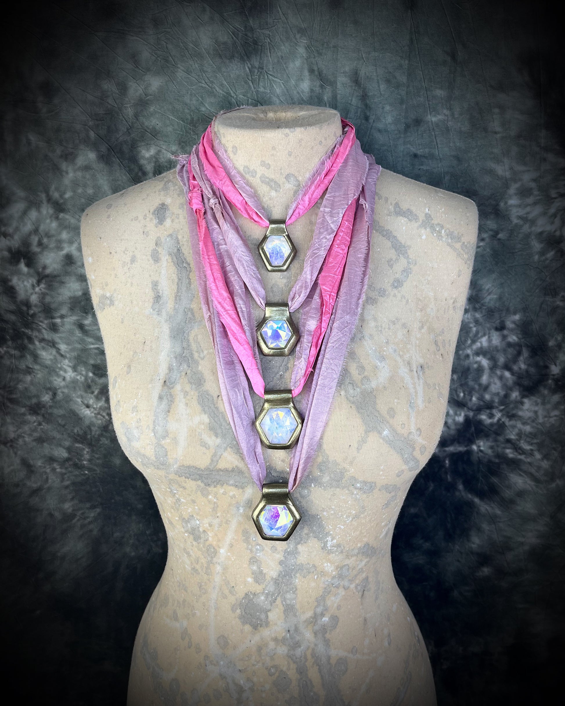 Shown here layered on a mannequin, handmade designer clear opalescent quartz gemstone layering pendants in a gold finish.  These crystal healing pyramid hexagon necklaces are strung from lilac and pink recycled silk for the sustainable and eco-friendly jewelry collector. 