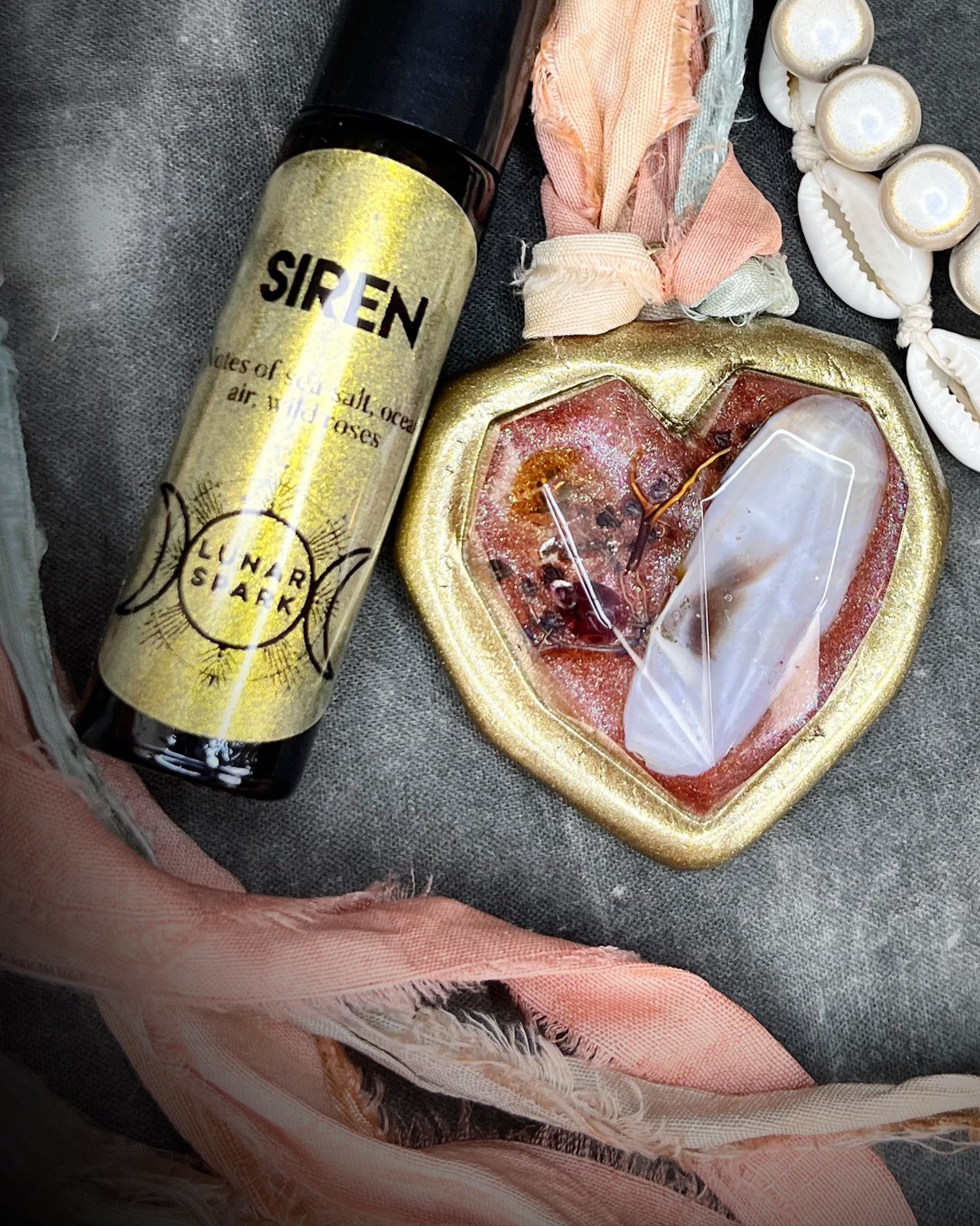 Gold bottle labeled 'Siren' with a heart-shaped crystal pendant and shell necklace on a textured surface.