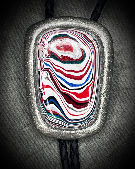 A close-up of a silver-toned bolo tie pendant featuring a marbled, swirled pattern of red, white, blue, and black glass, suspended on a black braided cord against a dark textured background.
