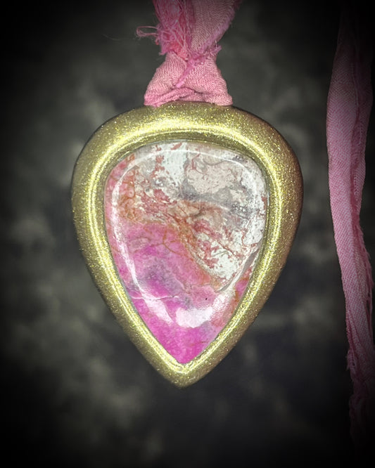 A teardrop-shaped pendant with a gold  border and a pink and white marbled pink calcite stone inside, hanging from a pink recycled silk ribbon against a dark textured background.