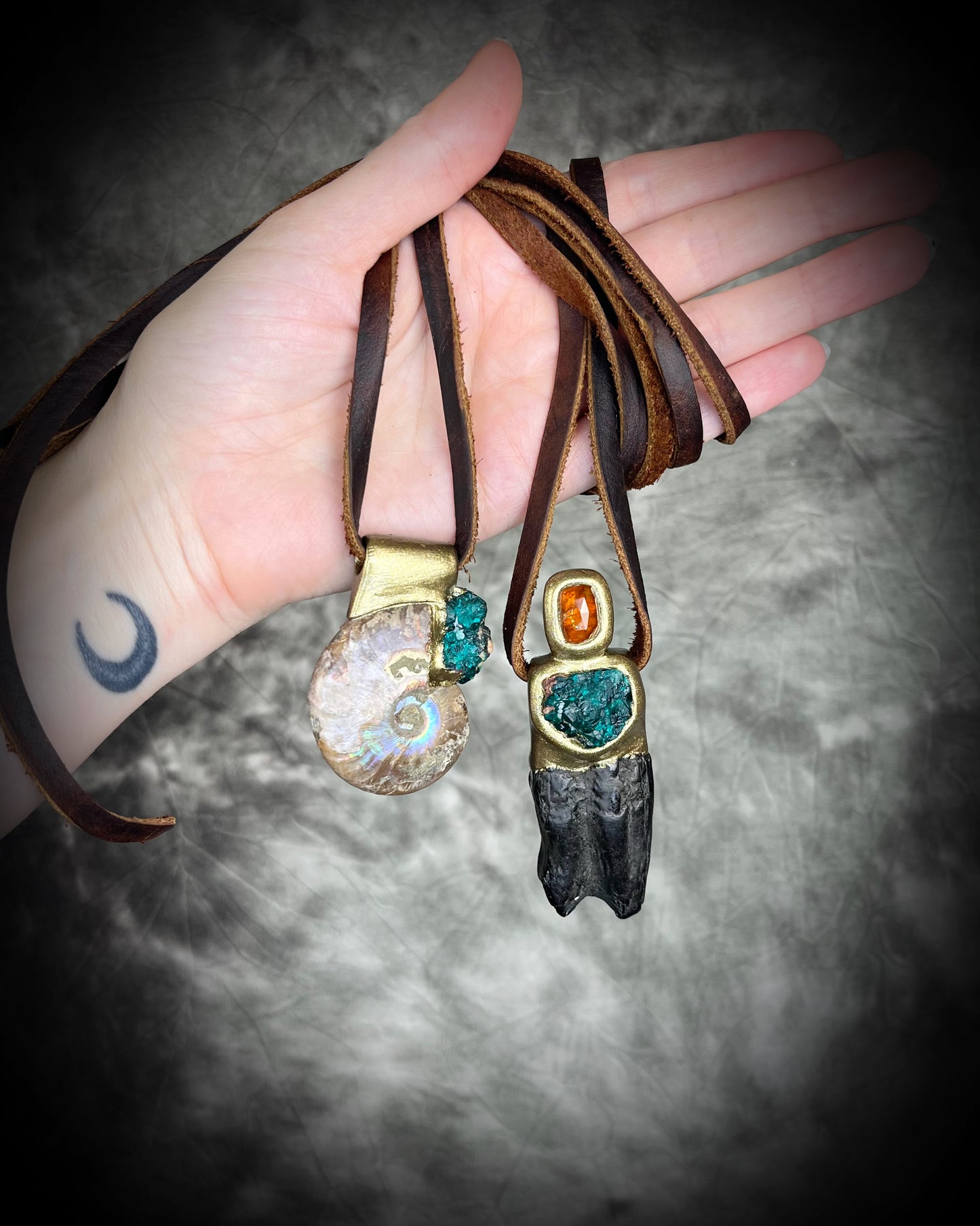 Ammonite fossil necklace with green dioptase crystals and Equus Horse tooth fossil necklace with orange kyanite gemstones. Both pendants are strung from earthy brown leather and held in a hand for display- best gift her for 2025 fall maximalism and spiritual jewelry trends. 