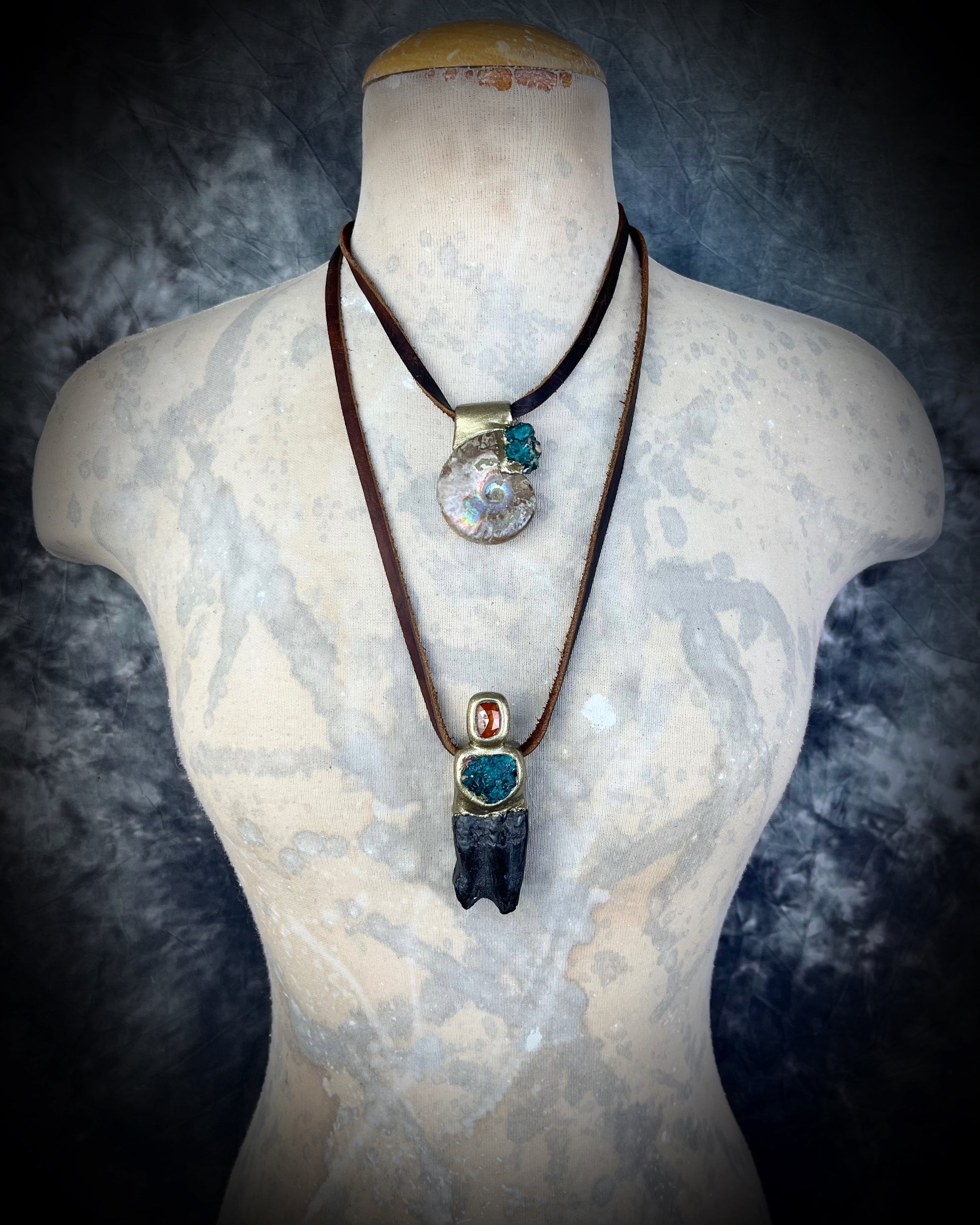 Ammonite fossil necklace with green dioptase crystals and Equus Horse tooth fossil necklace with orange kyanite gemstones. Both pendants are strung from earthy brown leather and layered for 2025 fall maximalism and spiritual jewelry trends. 