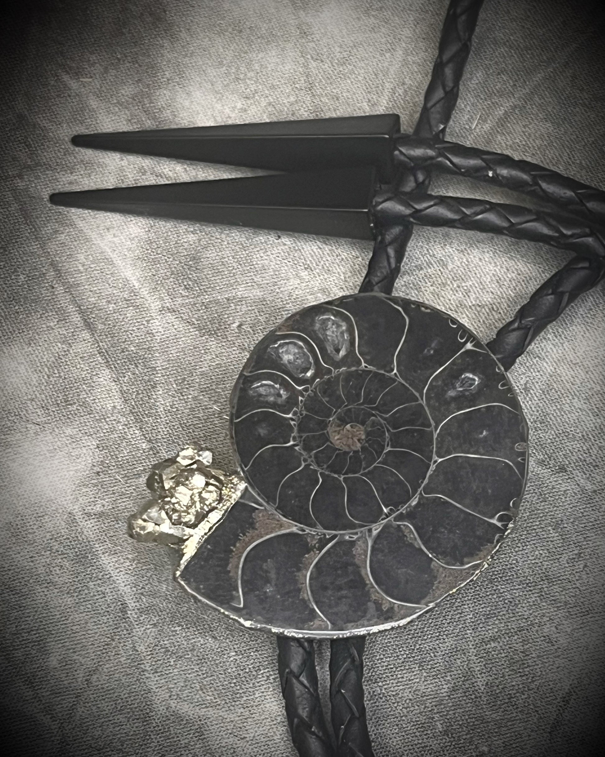 Black ammonite fossil and pyrite bolo tie for men on black leather with black spike tips, Western gothic statement jewelry with fossil and crystal detail, handmade in Baltimore Maryland.