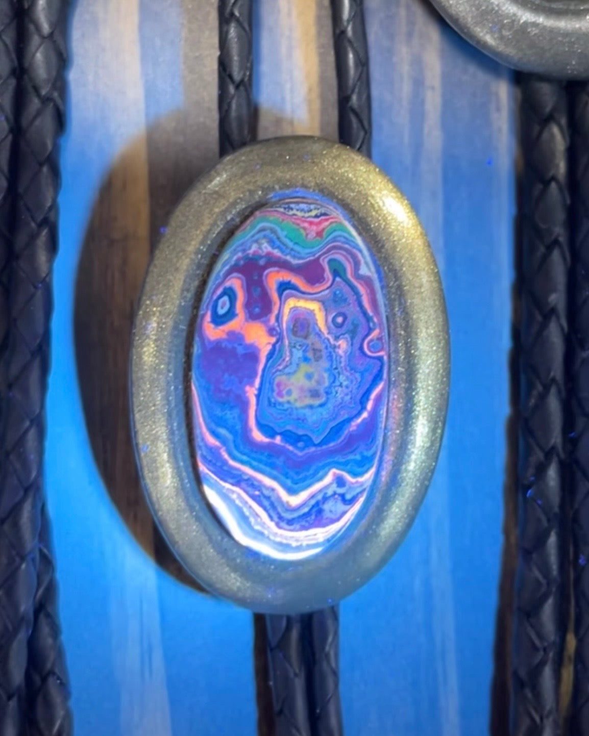 Cadillac Ranch Fordite manmade gemstone UV reactive Bolo tie set in a gold finish and mounted on soft black bolo leather. A talisman for the modern man, crystal jewelry for him. Western 2026 jewelry trend bolo. 