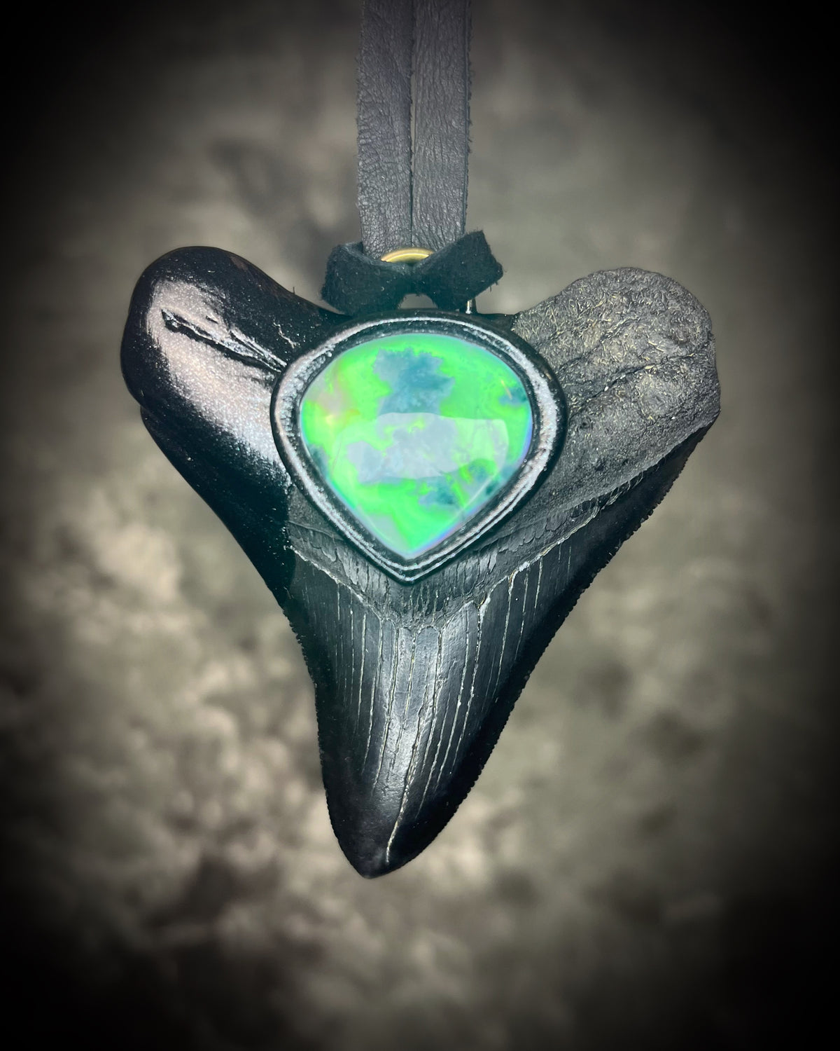 Black megalodon tooth talisman with a glowing green moss agate teardrop focal stone in a sculptural black-finish setting on black leather, handmade by Become Spellbound in Baltimore, Maryland.
