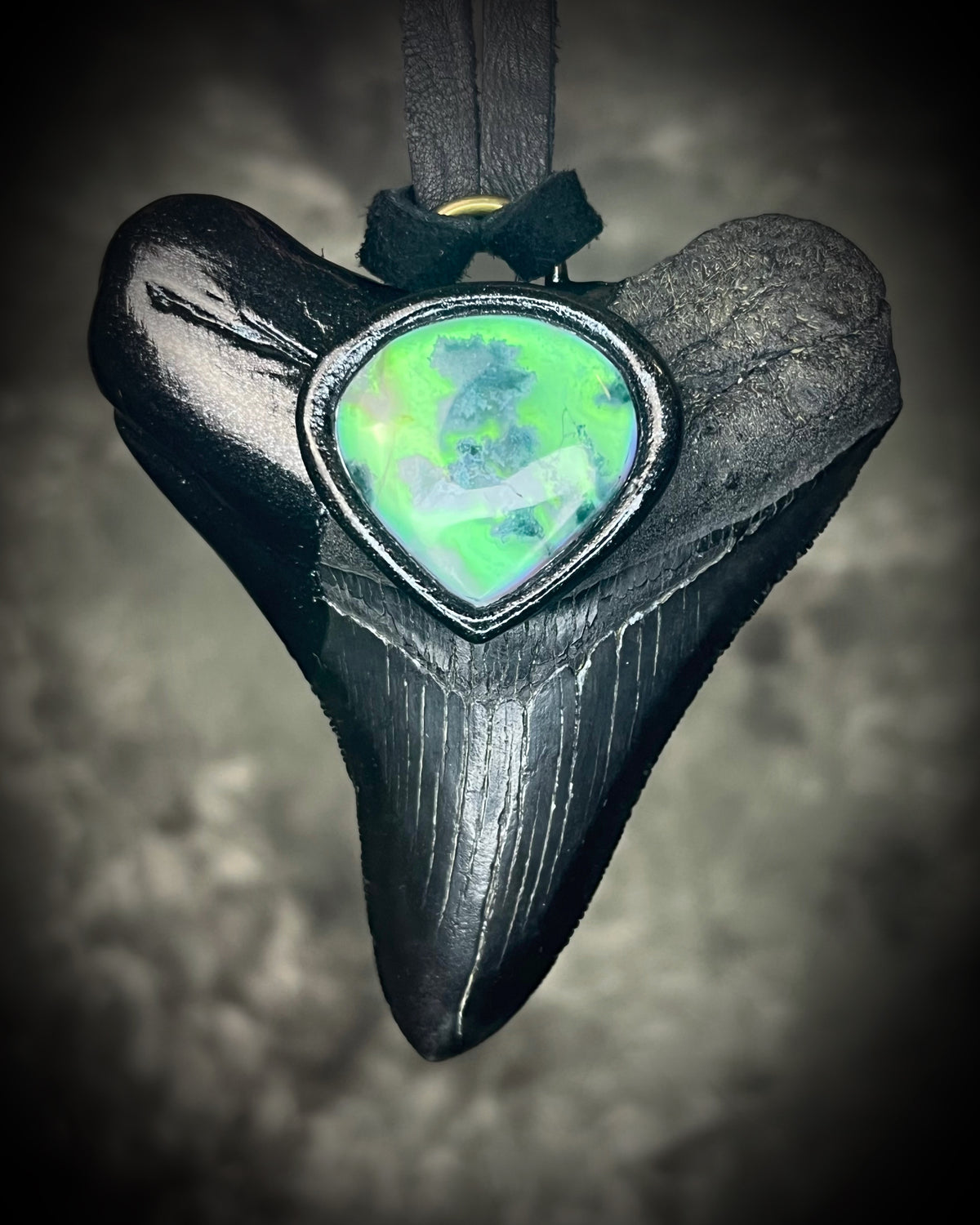Black megalodon tooth talisman with a glowing green moss agate teardrop focal stone in a sculptural black-finish setting on black leather, handmade by Become Spellbound in Baltimore, Maryland.