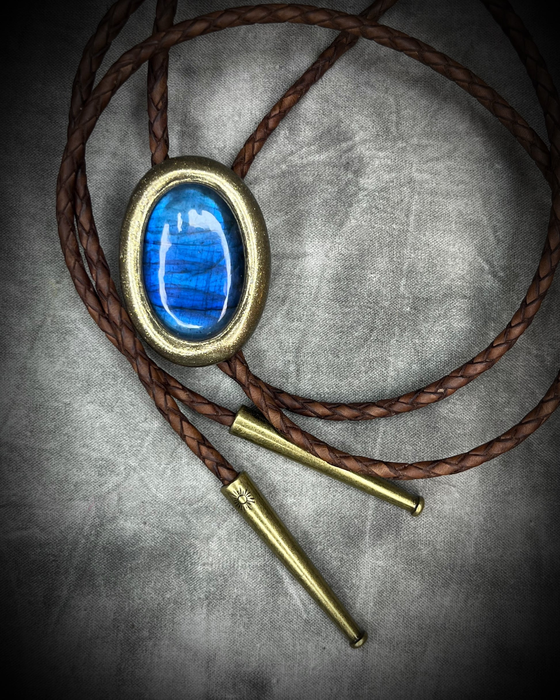 Blue flash labradorite bolo tie with brown leather cord, gold finish, and decorative sunburst bolo tips. Handmade by Become Spellbound.