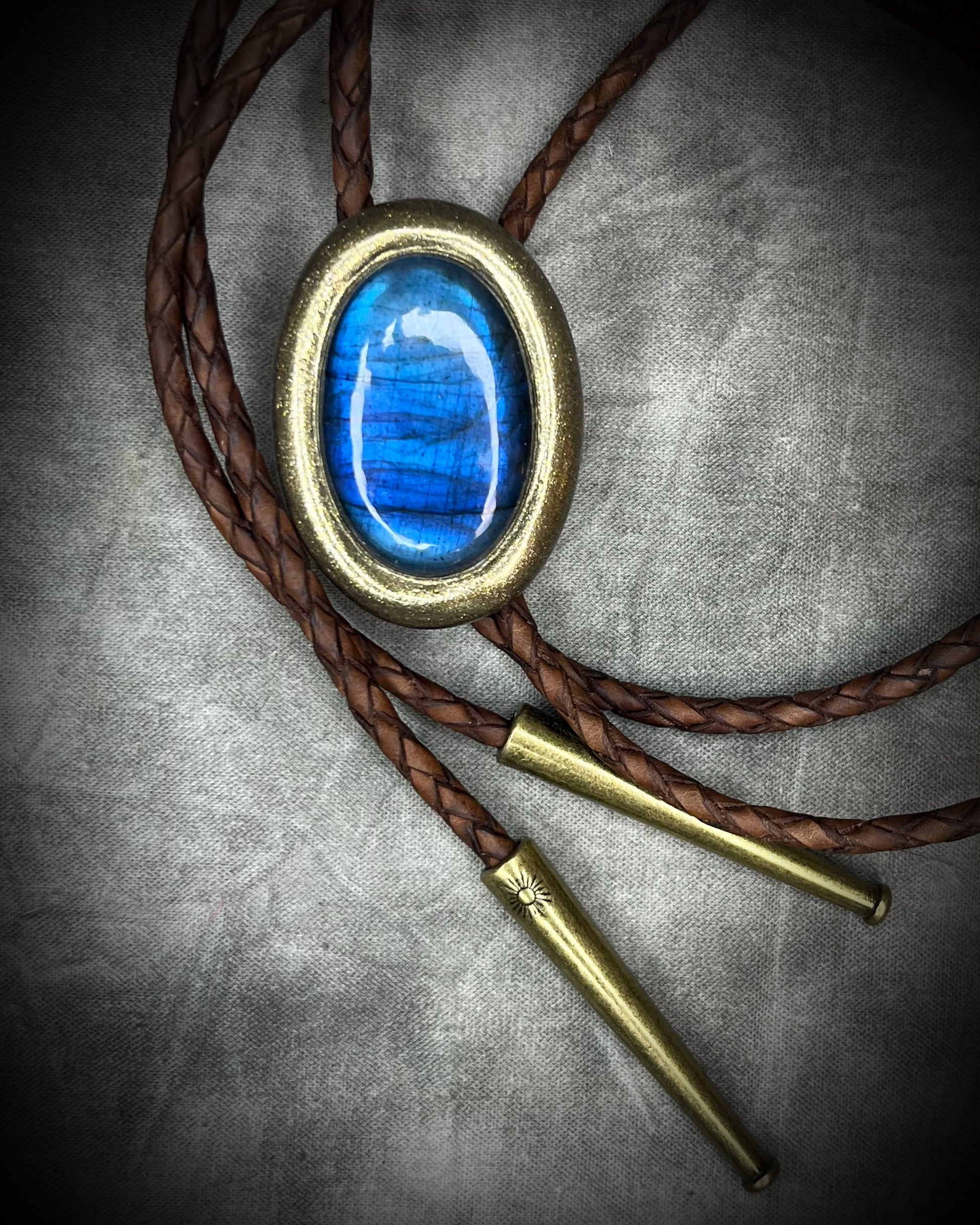 Blue flash labradorite bolo tie with brown leather cord, gold finish, and decorative sunburst bolo tips. Handmade by Become Spellbound.