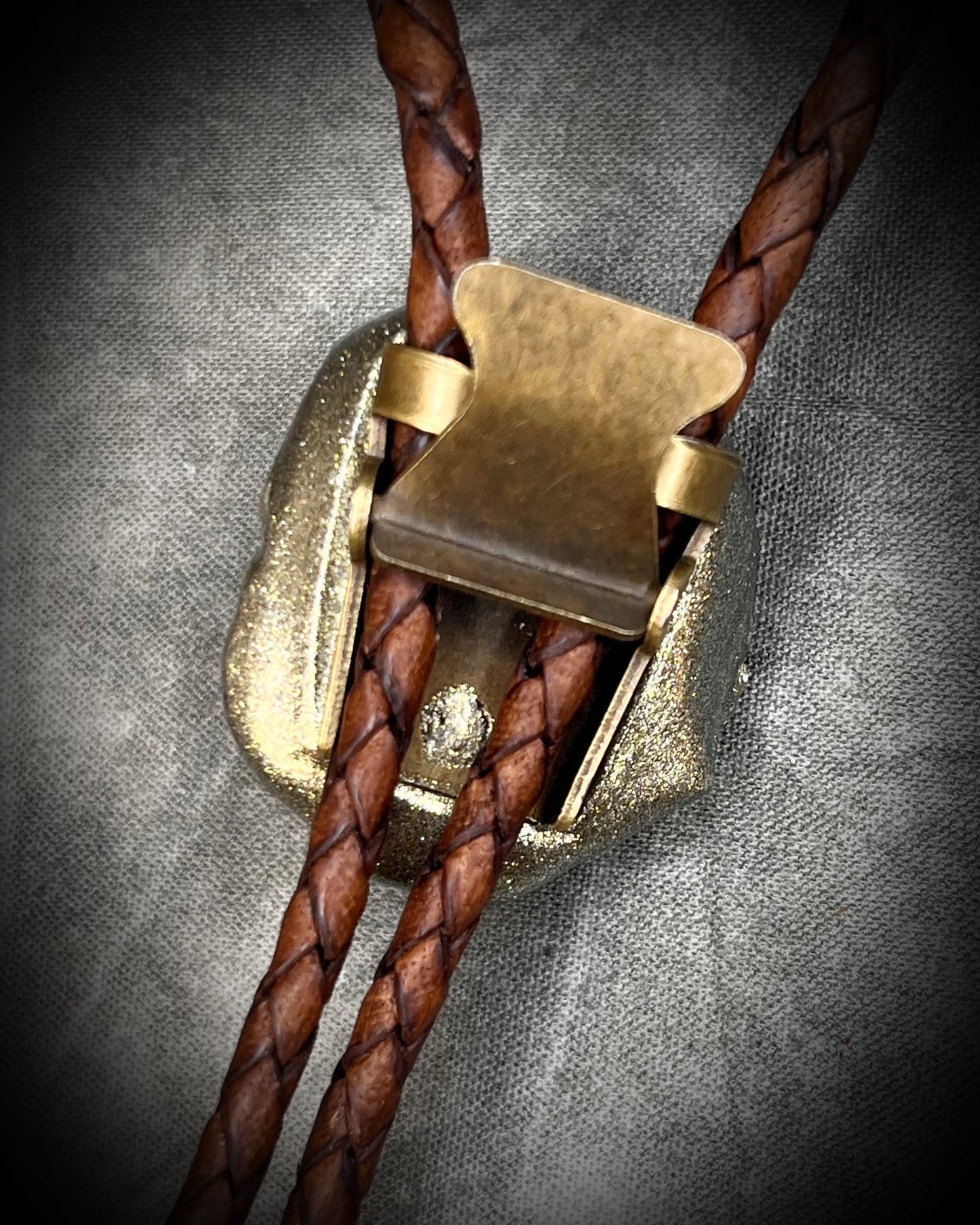 Close-up of the back clasp of a bolo tie with braided brown leather and brass clasp on a textured gray background