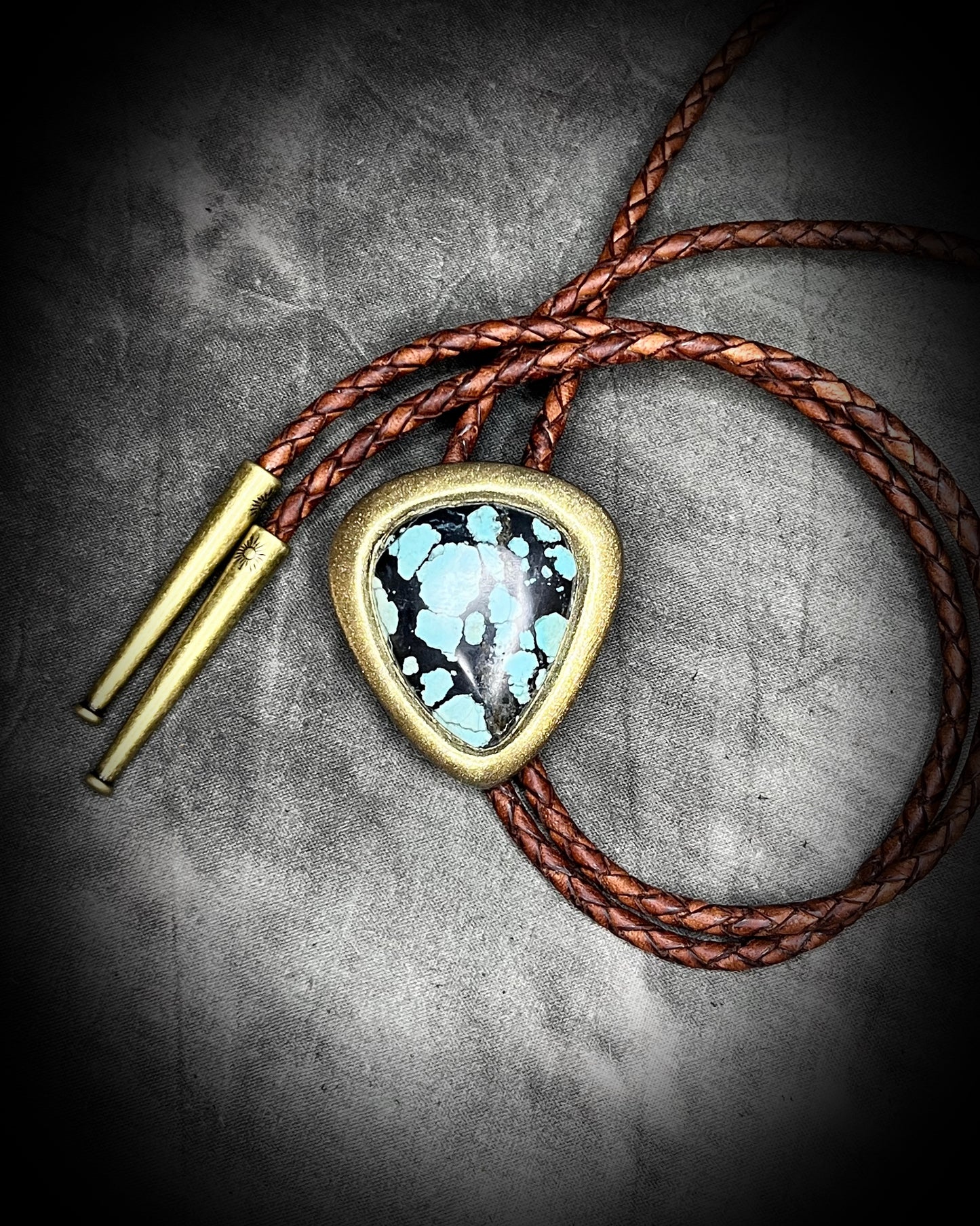 Bolo tie with turquoise stone, brown leather and gold accents on a textured gray background