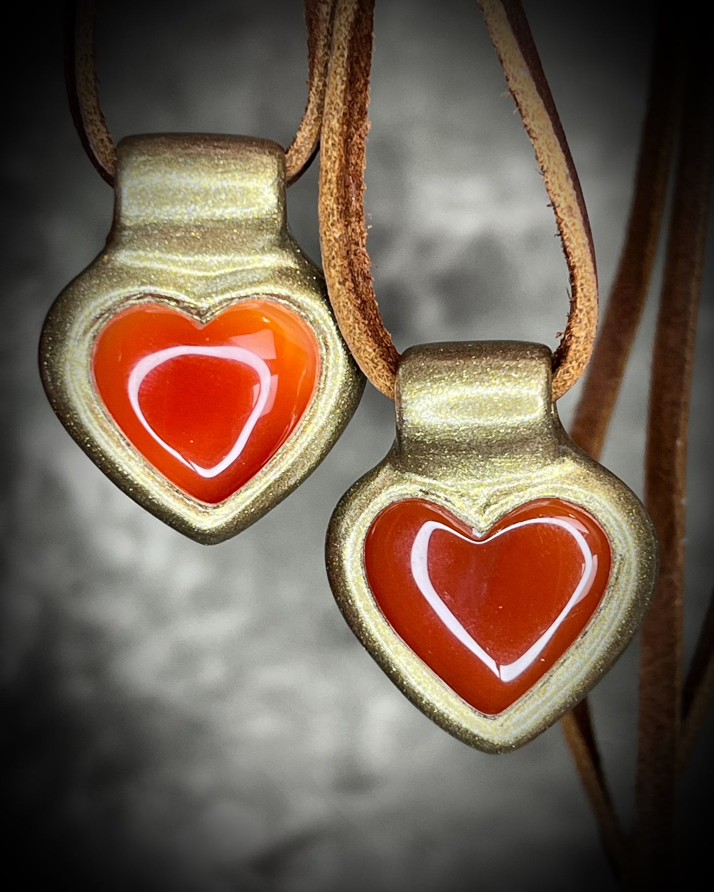 Heart-shaped pendants with red hearts and gold frames on a leather cord against a dark background.