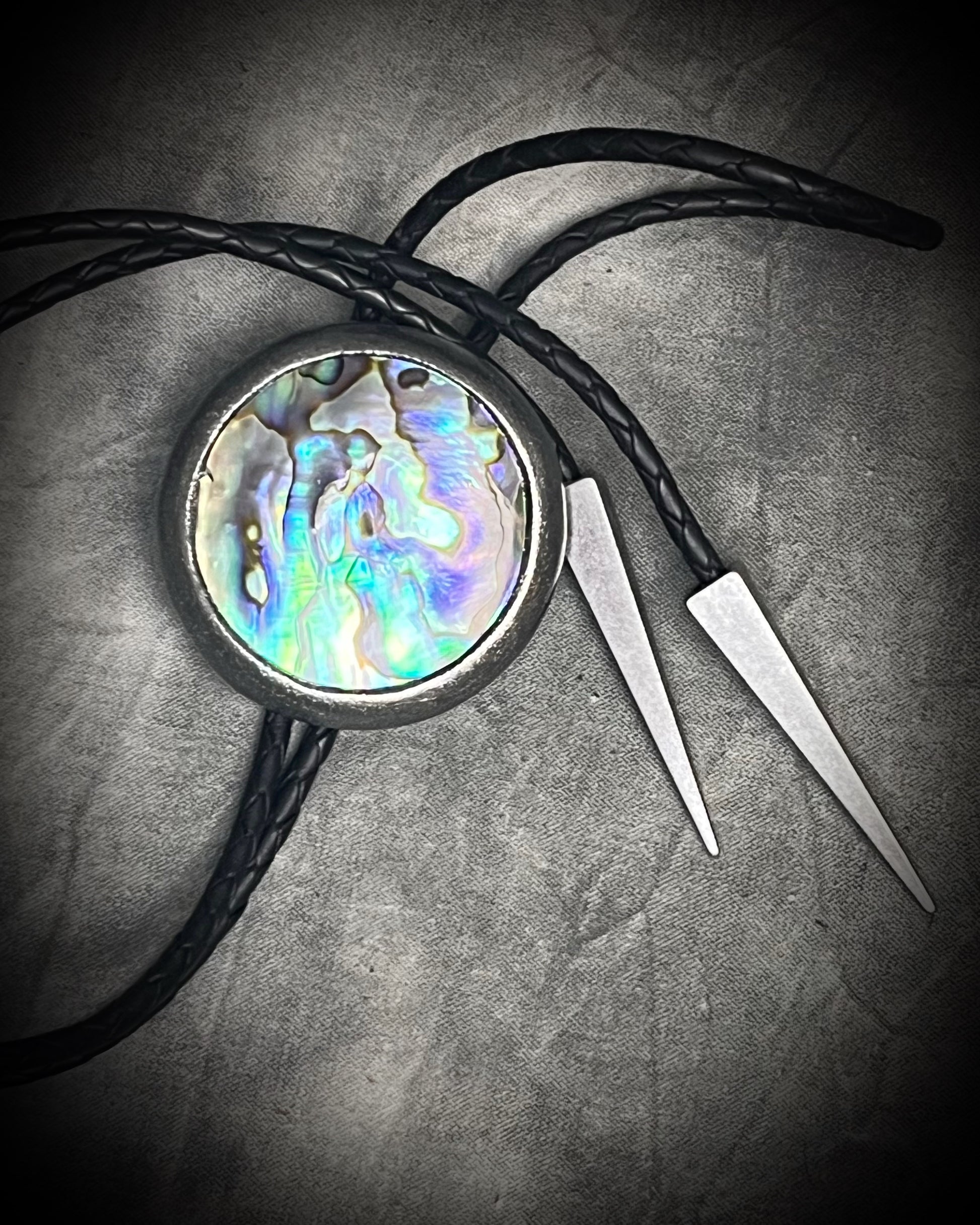 Circular abalone shell bolo tie with blue, green, and lilac iridescence, dark silver setting, black leather cord, and gunmetal silver bolo tips. Handmade by Become Spellbound.