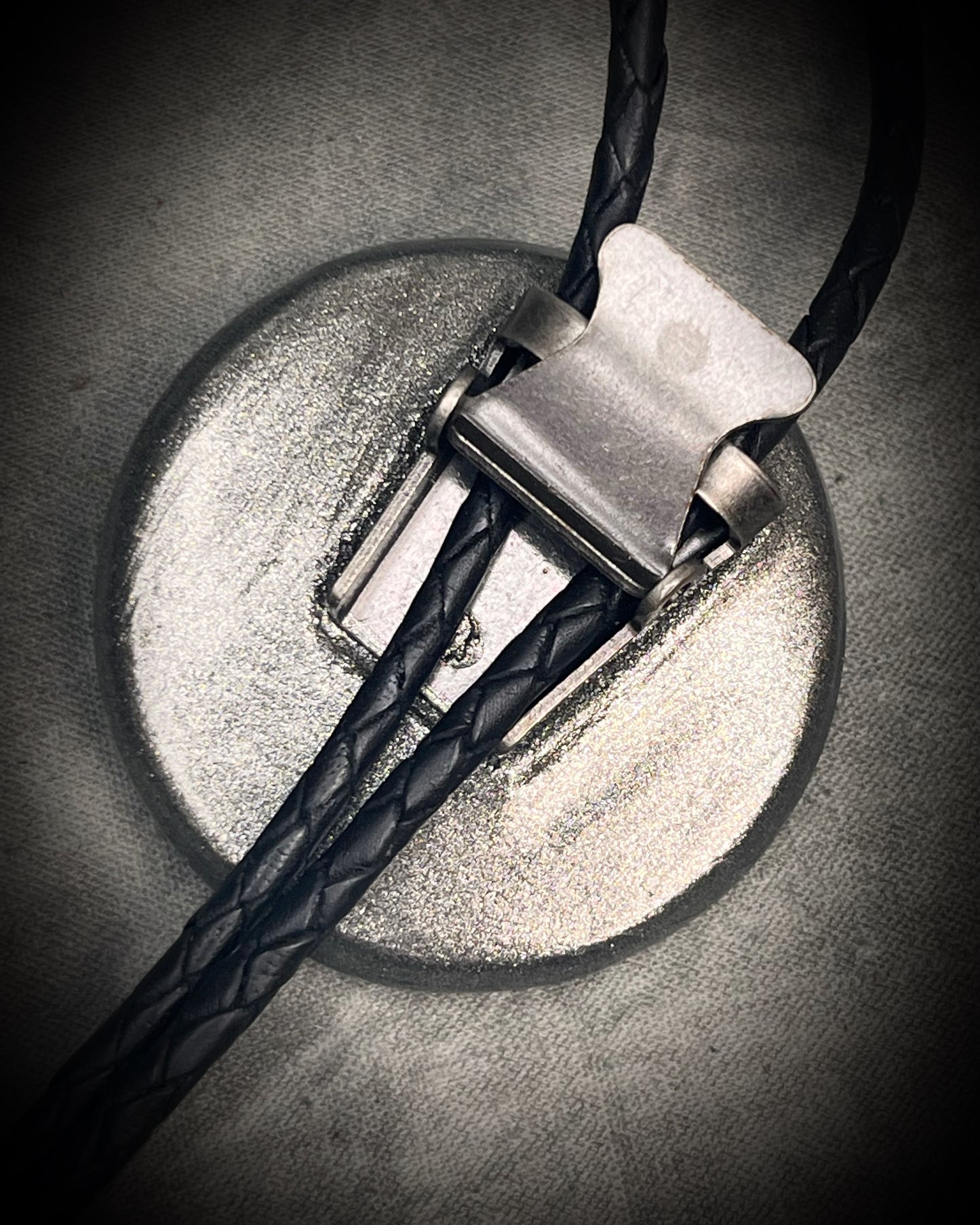 Circular abalone shell bolo tie with blue, green, and lilac iridescence, dark silver setting, black leather cord, and gunmetal silver bolo tips. Handmade by Become Spellbound.