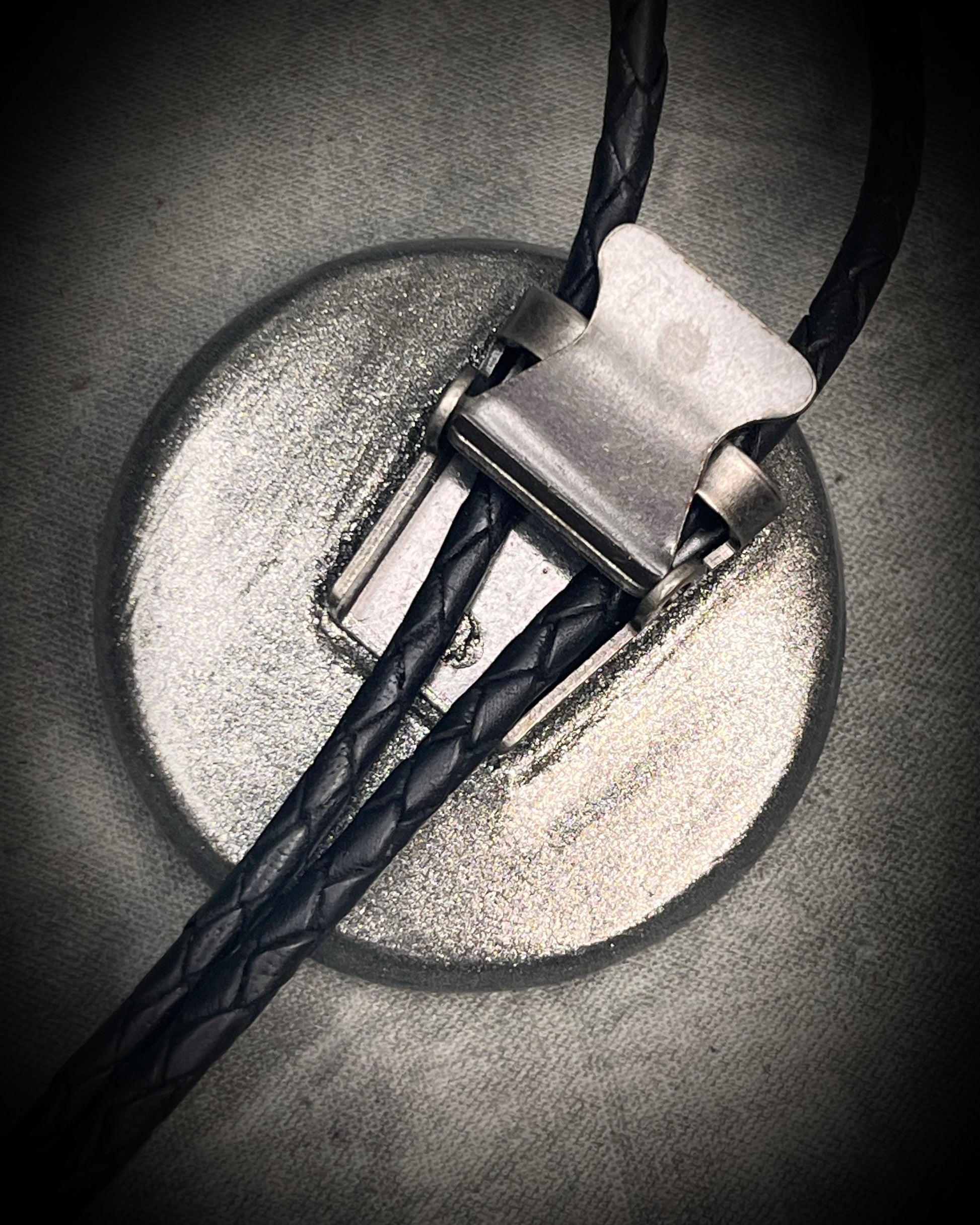 Circular abalone shell bolo tie with blue, green, and lilac iridescence, dark silver setting, black leather cord, and gunmetal silver bolo tips. Handmade by Become Spellbound.