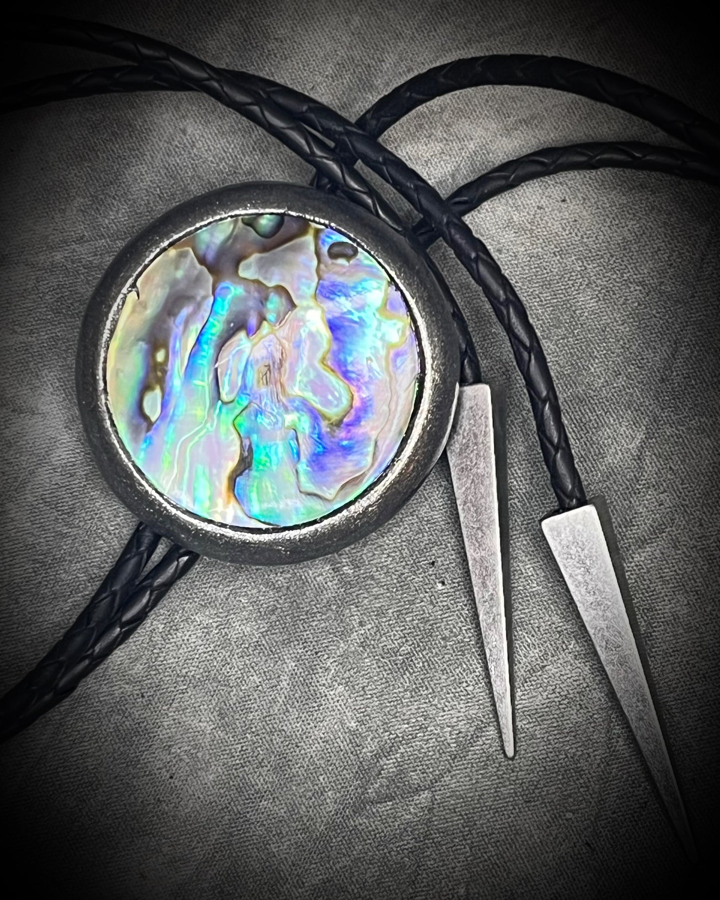 Circular abalone shell bolo tie with blue, green, and lilac iridescence, dark silver setting, black leather cord, and gunmetal silver bolo tips. Handmade by Become Spellbound.