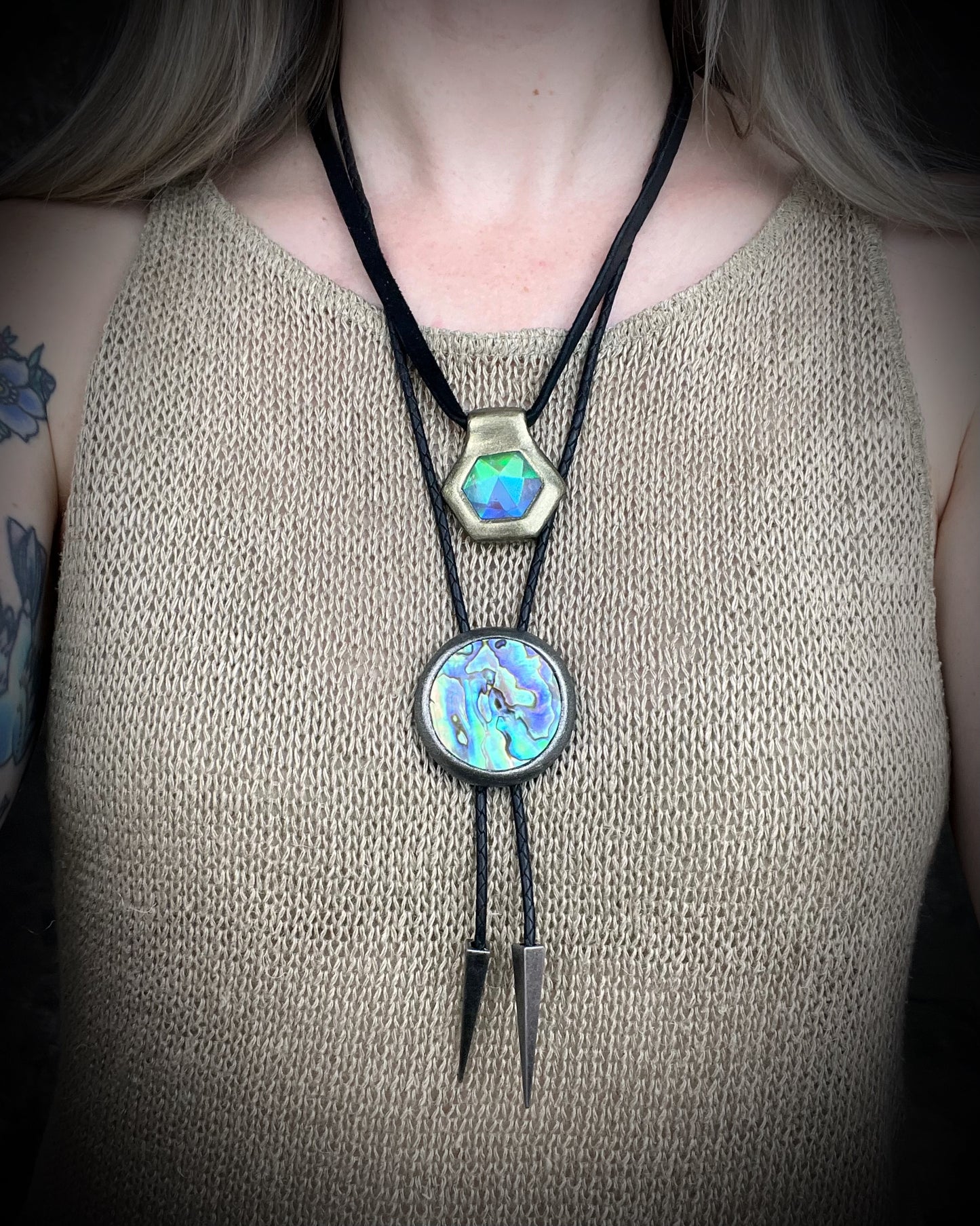 Circular abalone shell bolo tie with blue, green, and lilac iridescence, dark silver setting, black leather cord, and gunmetal silver bolo tips. Handmade by Become Spellbound.