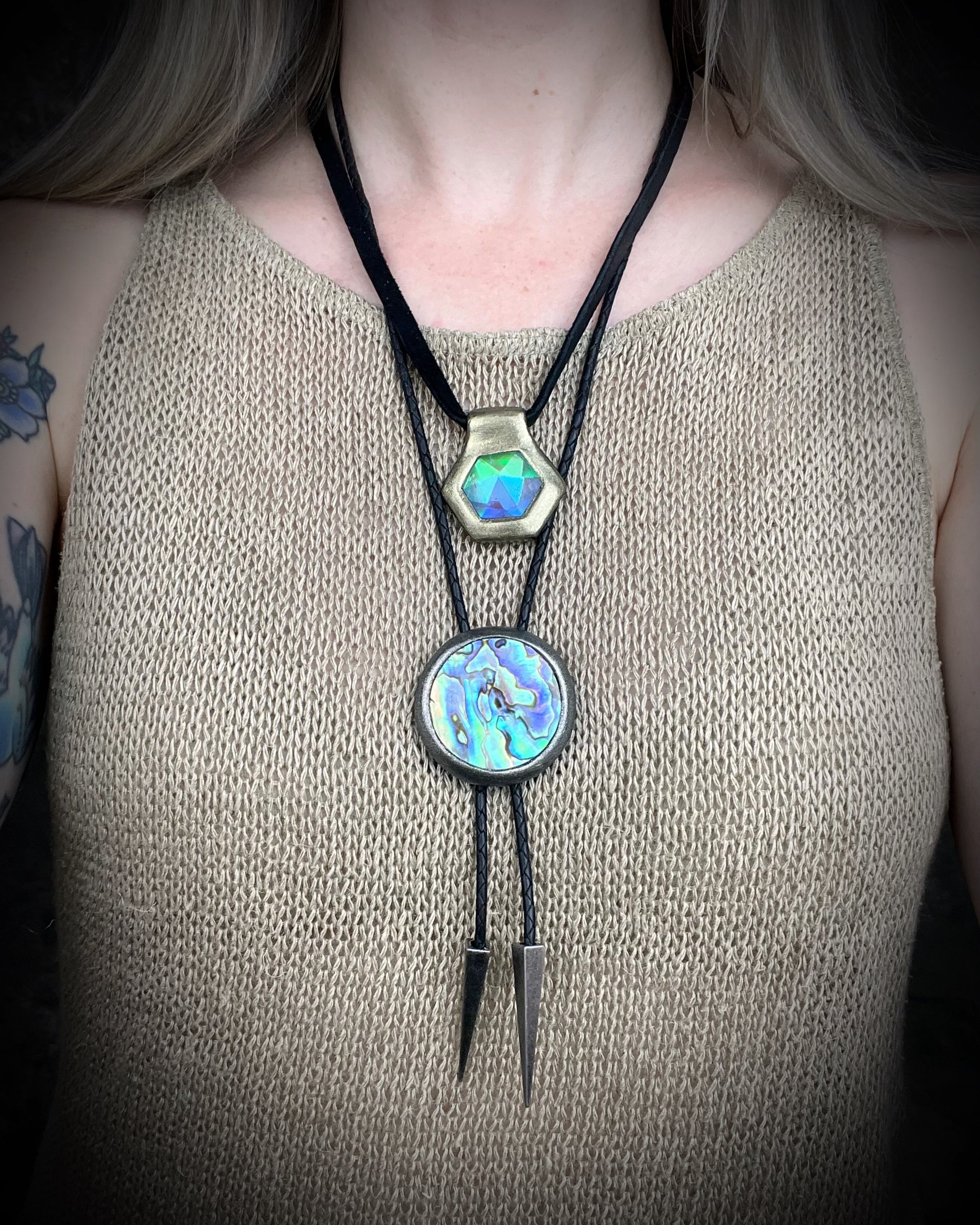Circular abalone shell bolo tie with blue, green, and lilac iridescence, dark silver setting, black leather cord, and gunmetal silver bolo tips. Handmade by Become Spellbound.
