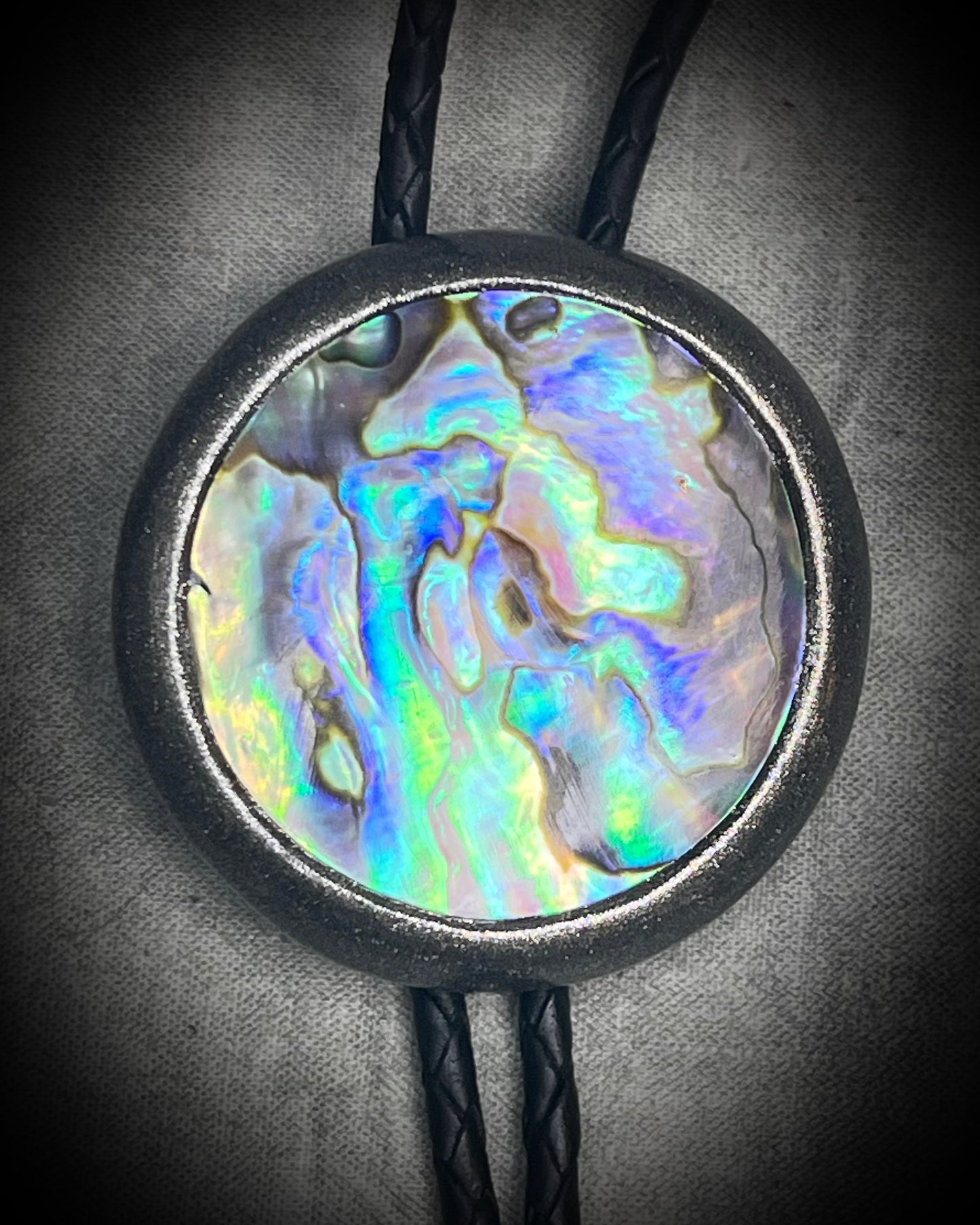 Circular abalone shell bolo tie with blue, green, and lilac iridescence, dark silver setting, black leather cord, and gunmetal silver bolo tips. Handmade by Become Spellbound.