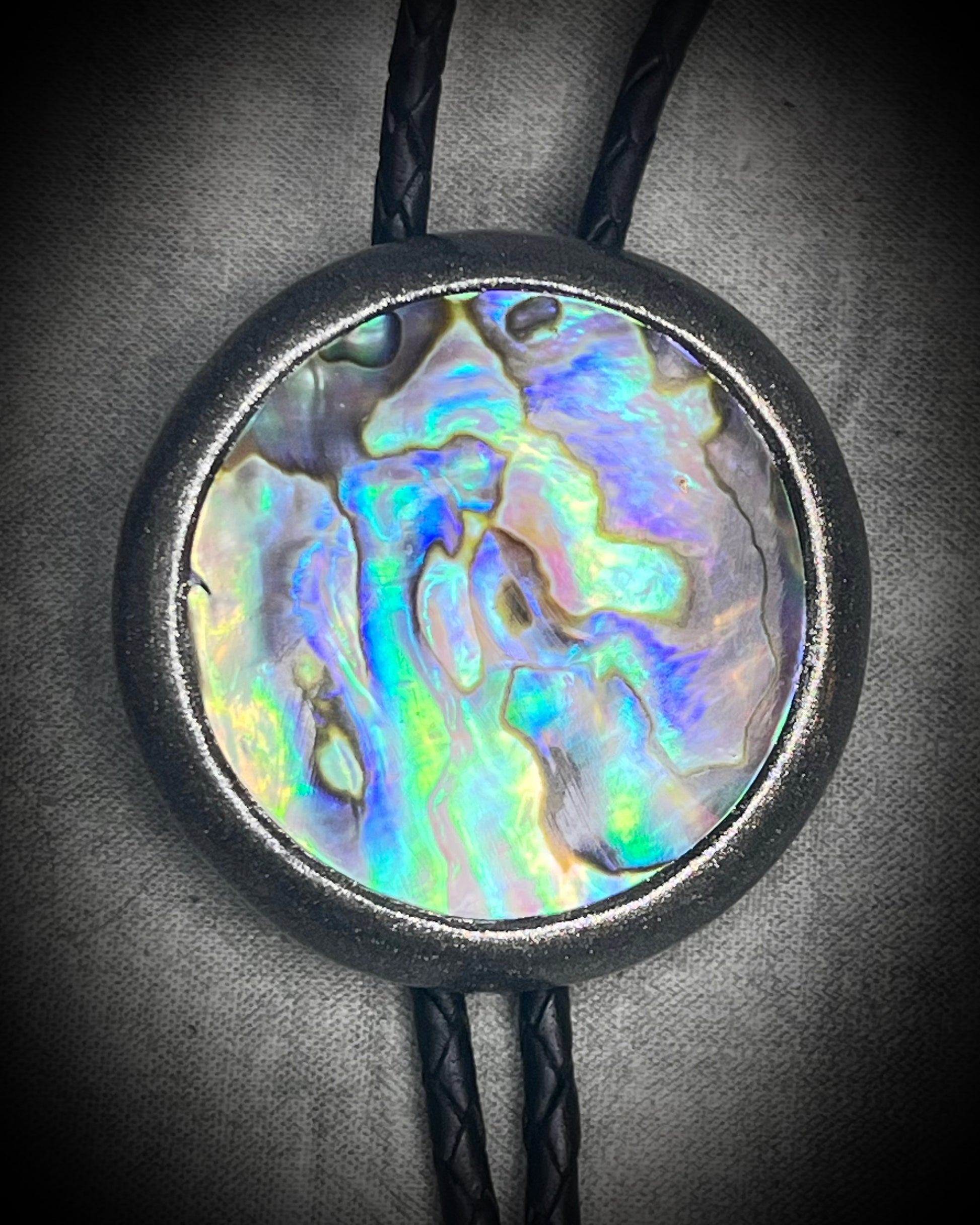 Circular abalone shell bolo tie with blue, green, and lilac iridescence, dark silver setting, black leather cord, and gunmetal silver bolo tips. Handmade by Become Spellbound.