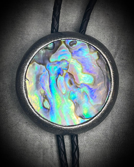Circular abalone shell bolo tie with blue, green, and lilac iridescence, dark silver setting, black leather cord, and gunmetal silver bolo tips. Handmade by Become Spellbound.