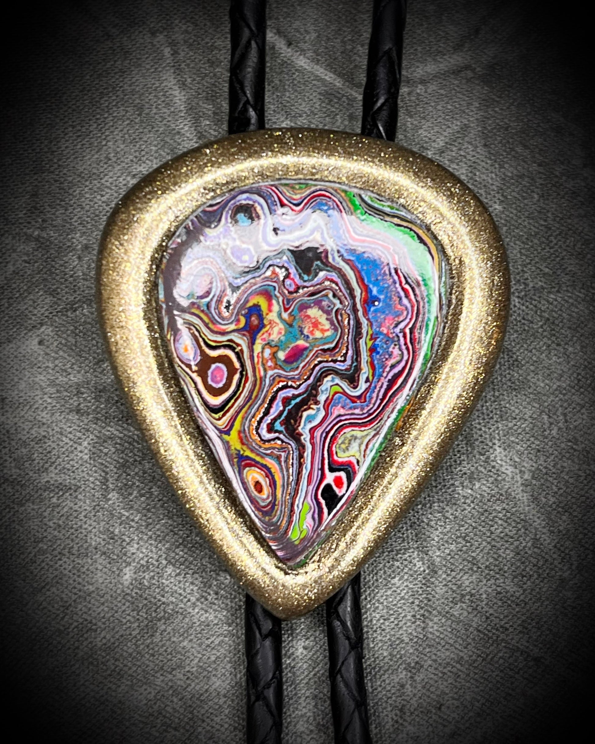 Closeup-Cadillac Ranch Fordite manmade colorful swirling gemstone Bolo tie set in a gold finish and mounted on soft black bolo leather. A talisman for the modern man, crystal jewelry for him. Western 2026 jewelry trend bolo. 