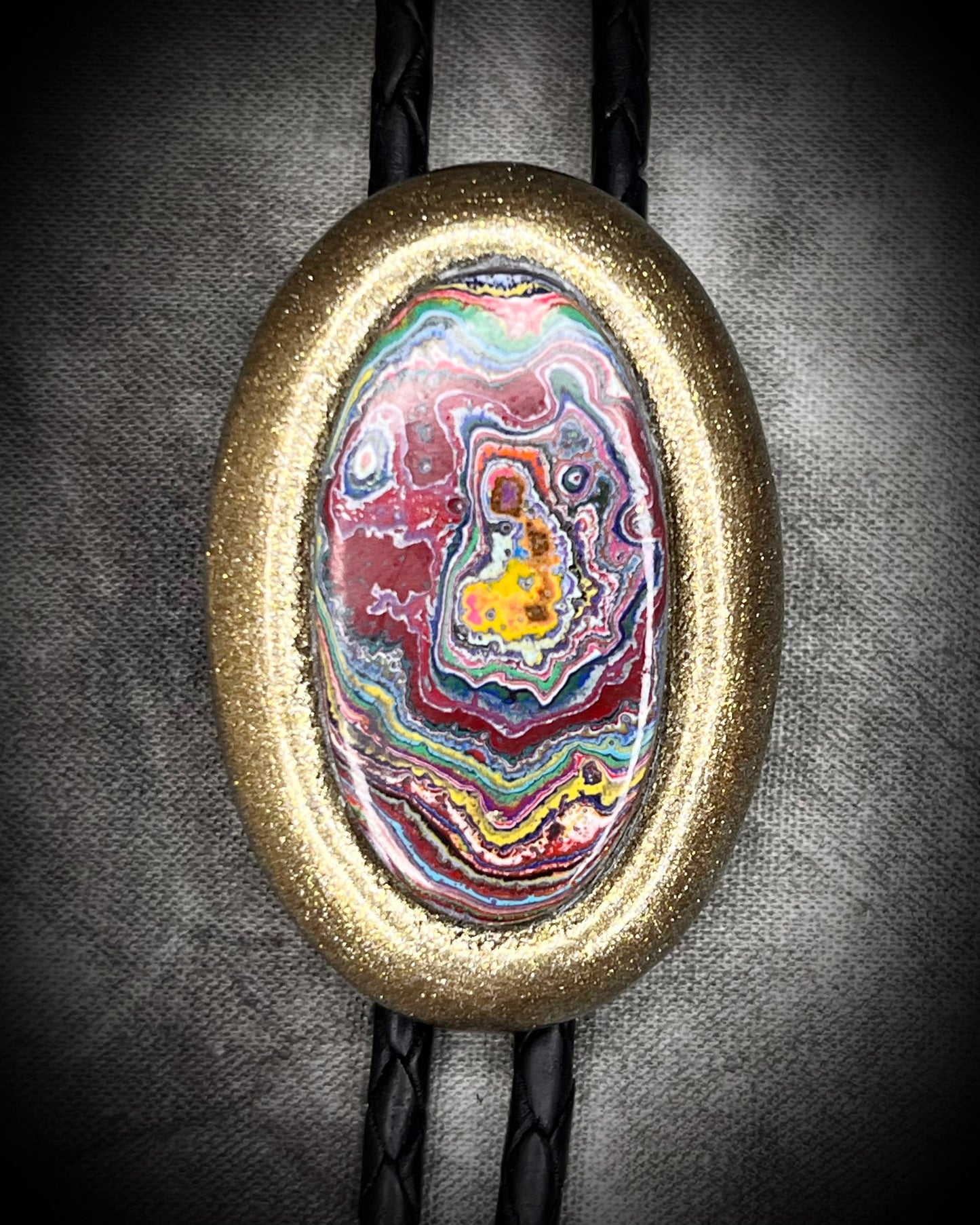 Closeup of Cadillac Ranch Fordite manmade colorful swirling gemstone Bolo tie set in a gold finish and mounted on soft black bolo leather. A talisman for the modern man, crystal jewelry for him. Western 2026 jewelry trend bolo. 