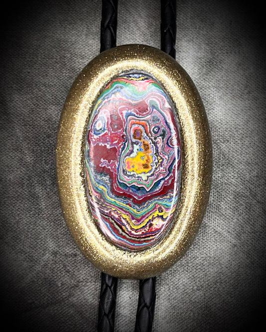 Closeup of Cadillac Ranch Fordite manmade colorful swirling gemstone Bolo tie set in a gold finish and mounted on soft black bolo leather. A talisman for the modern man, crystal jewelry for him. Western 2026 jewelry trend bolo. 