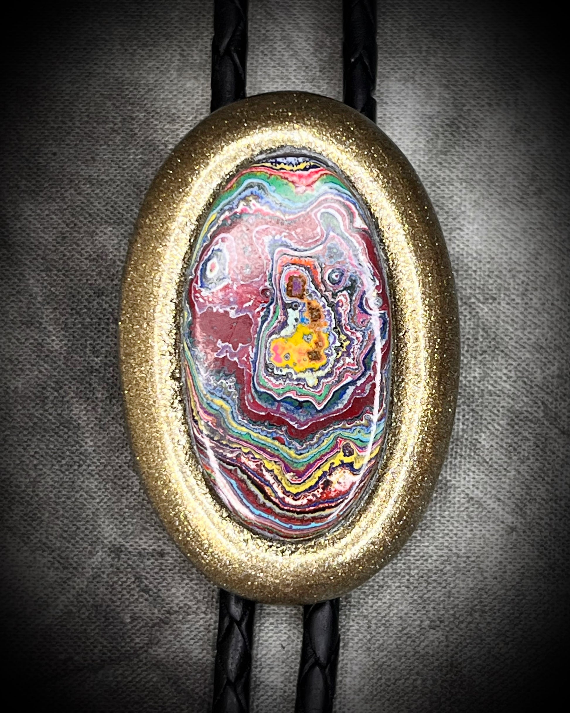 Closeup of Cadillac Ranch Fordite manmade colorful swirling gemstone Bolo tie set in a gold finish and mounted on soft black bolo leather. A talisman for the modern man, crystal jewelry for him. Western 2026 jewelry trend bolo. 