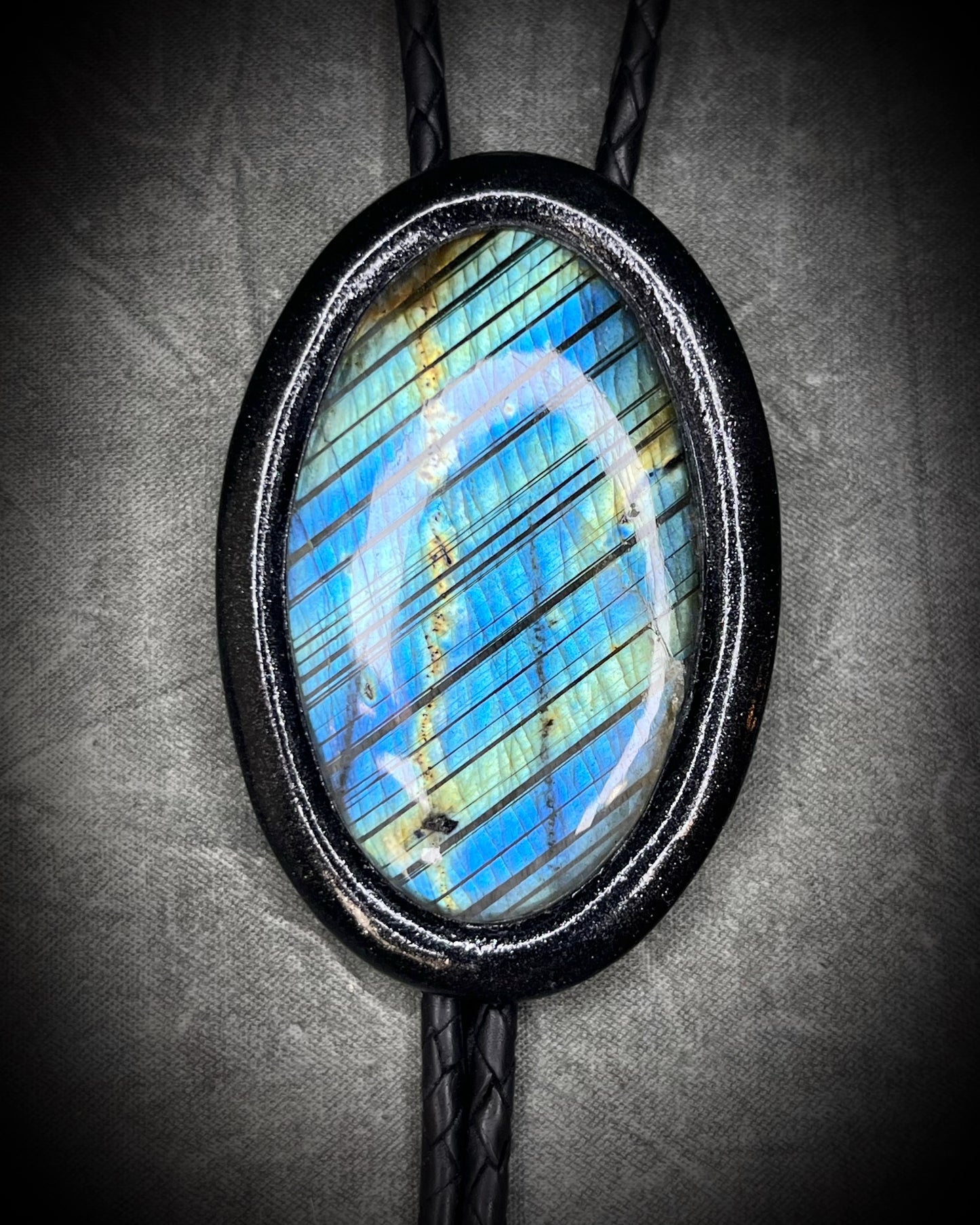Closeup of flashy stone on Handmade Bolo Tie Necklace with a blue and green labradorite pendant in black frame and leather cord on a textured gray background. 2026 Gothic western bolo trend in men's jewelry.
