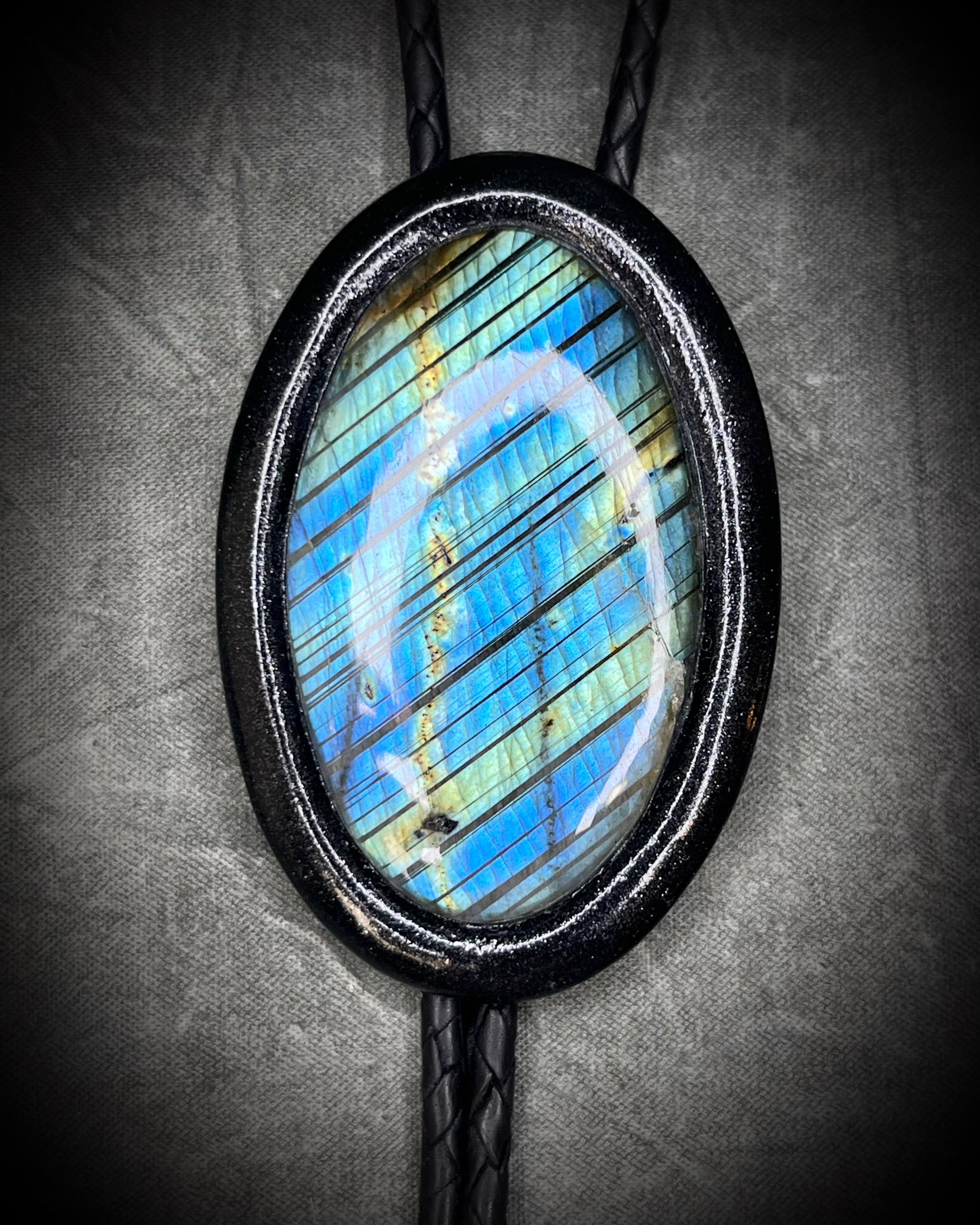 Closeup of flashy stone on Handmade Bolo Tie Necklace with a blue and green labradorite pendant in black frame and leather cord on a textured gray background. 2026 Gothic western bolo trend in men's jewelry.