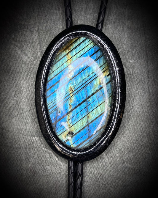 Closeup of flashy stone on Handmade Bolo Tie Necklace with a blue and green labradorite pendant in black frame and leather cord on a textured gray background. 2026 Gothic western bolo trend in men's jewelry.