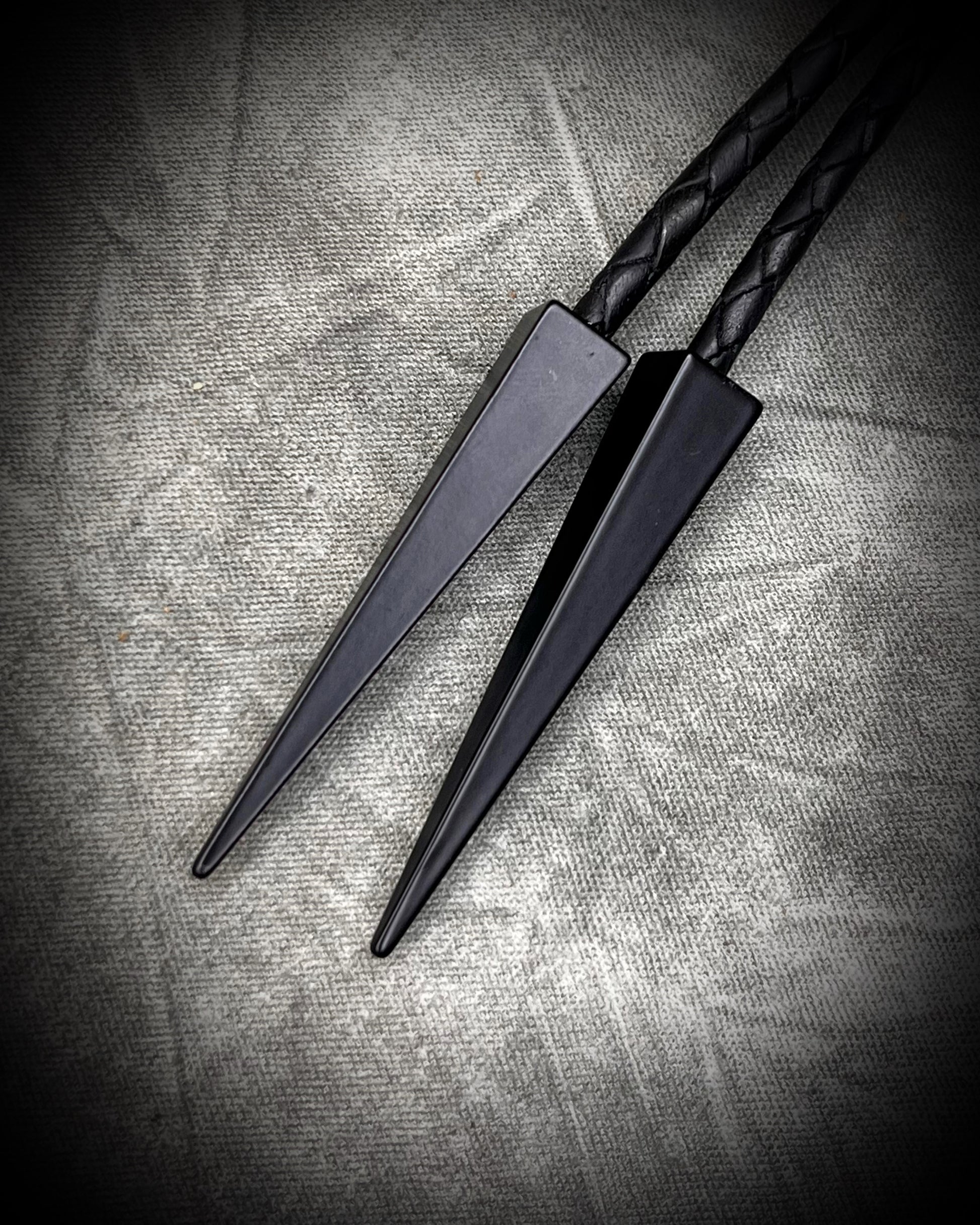 Closeup of matte black bolo tips  on Handmade Bolo Tie Necklace with a blue and green labradorite pendant in black frame and leather cord on a textured gray background. 2026 Gothic western bolo trend in men's jewelry.