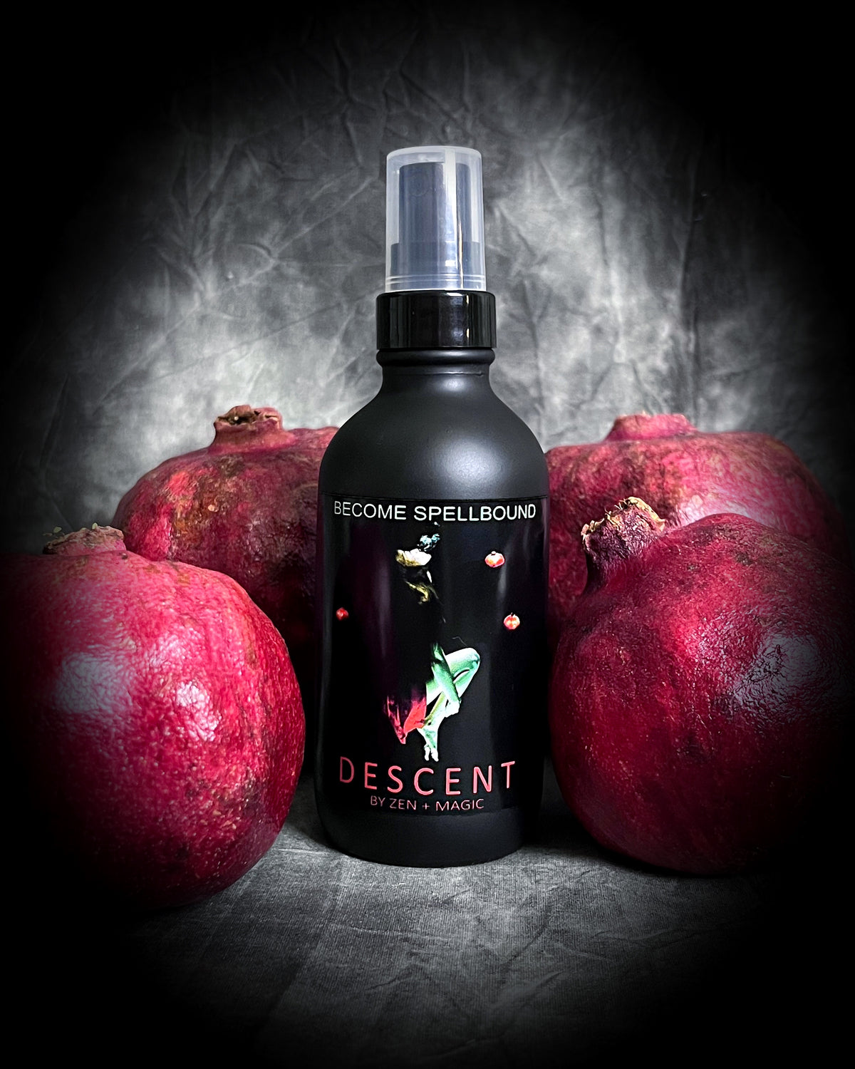 A beautiful matte black perfume bottle with a label that depicts a girl in a red dress going underwater and blowing out bubbles, surrounded by pomegranates.  Descent is an energetic spray for manifesting your desires.