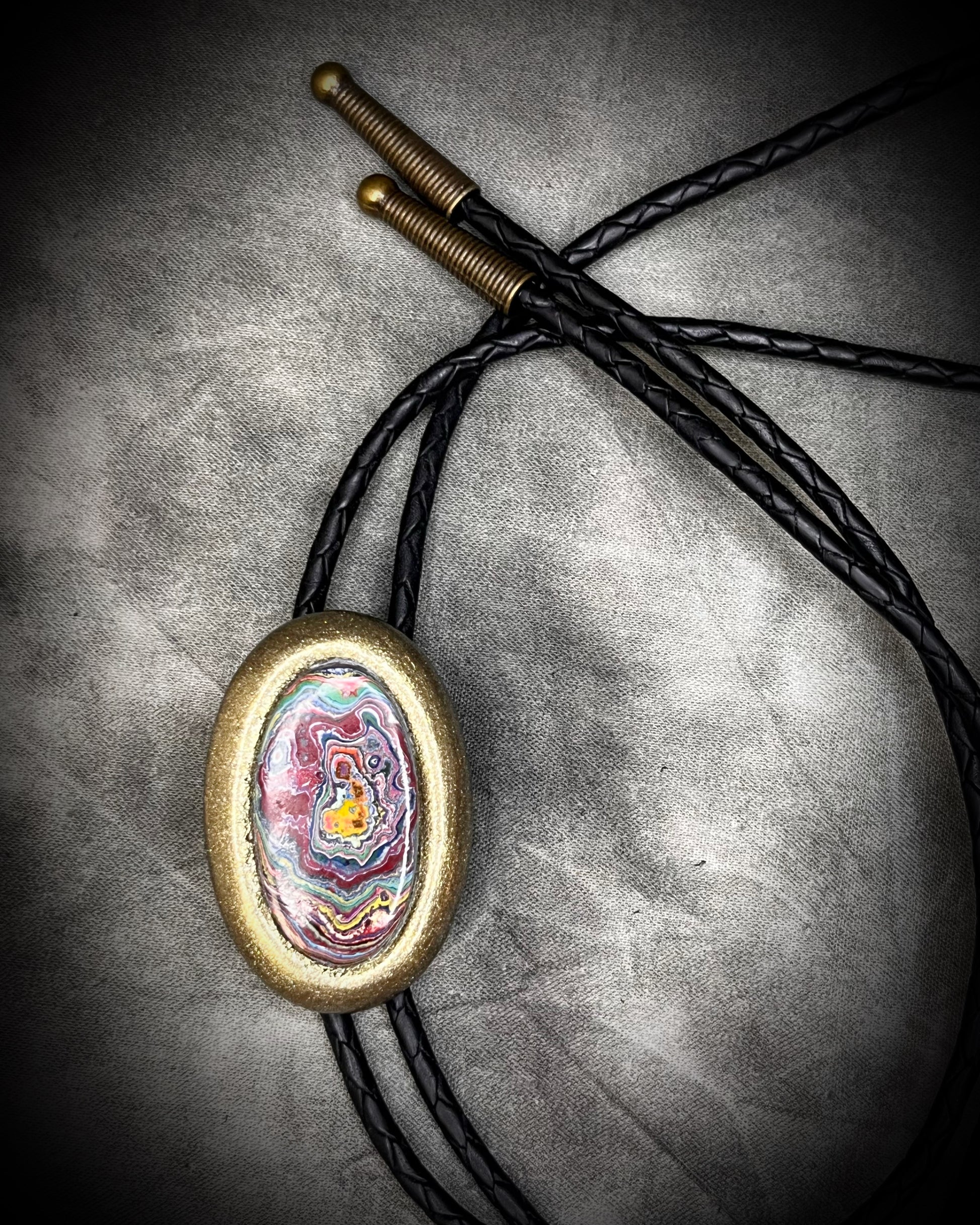 Flat lay of Cadillac Ranch Fordite manmade colorful swirling gemstone Bolo tie set in a gold finish and mounted on soft black bolo leather. A talisman for the modern man, crystal jewelry for him. Western 2026 jewelry trend bolo. 