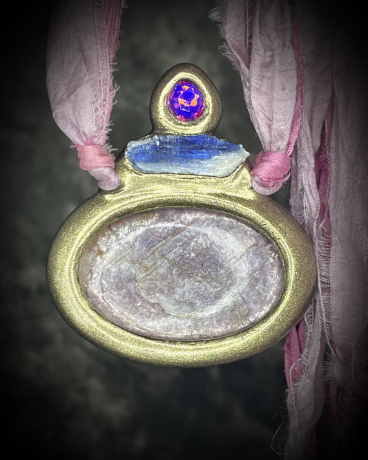 Gem lepidolite crystal talisman accented with blue kyanite and pink zircon in a sculptural gold-finish setting on pink recycled silk cords, handmade by Become Spellbound in Baltimore, Maryland.
