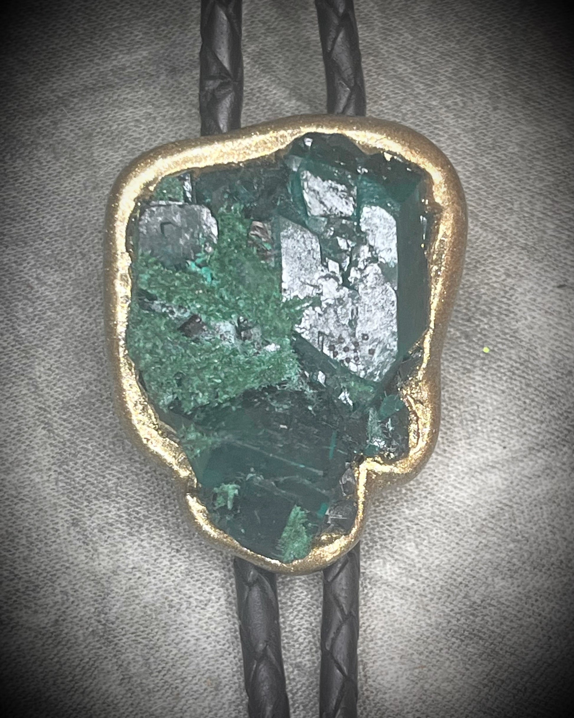 Gemmy Dioptase crystal bolo tie in deep green hues framed in a gold finish and strung on black bolo tie leather with gold tone brass spike tips. Gemstone bolo tie western inspired jewelry for him. Handmade in Baltimore, Maryland.