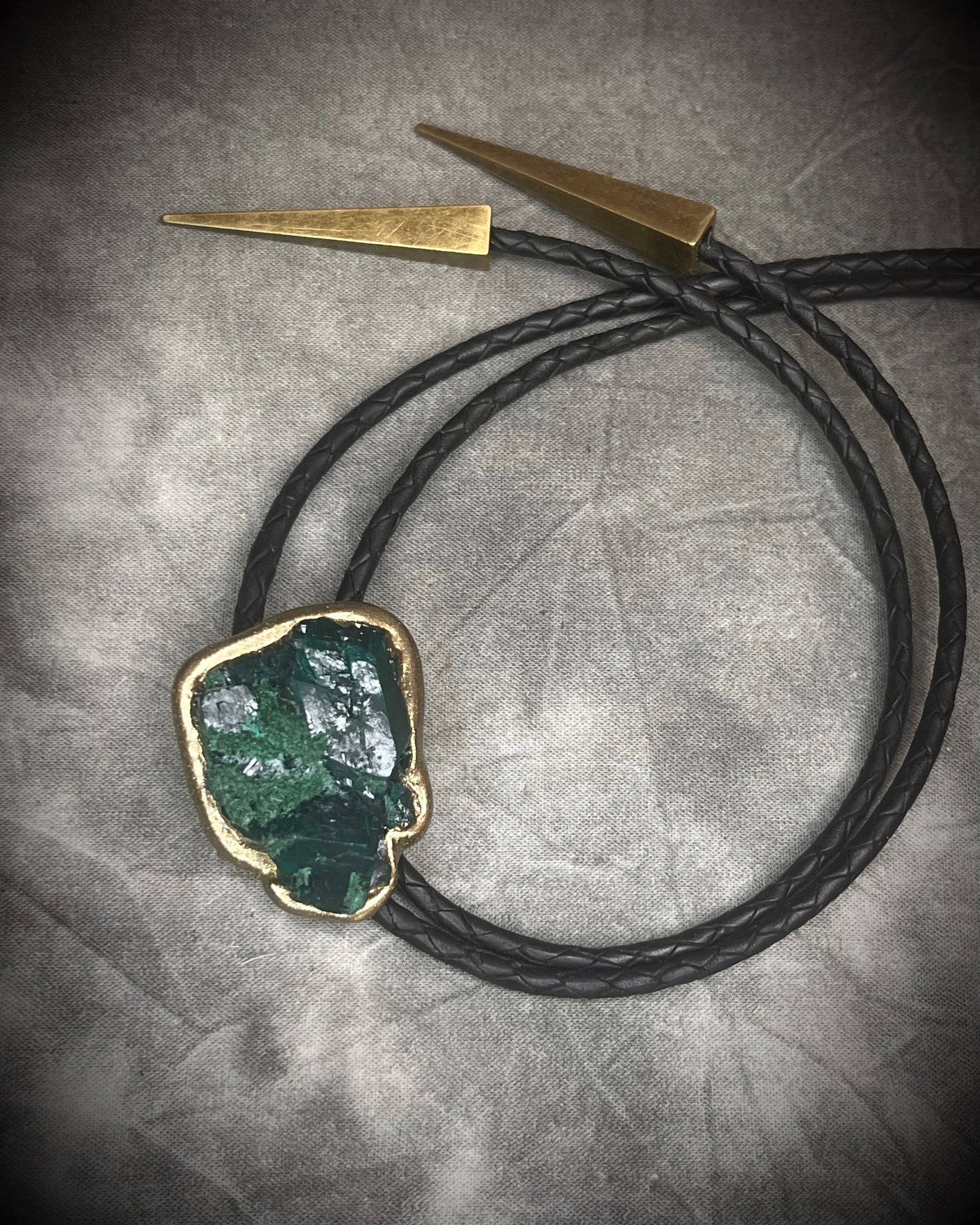 Gemmy Dioptase crystal bolo tie in deep green hues framed in a gold finish and strung on black bolo tie leather with gold tone brass spike tips. Gemstone bolo tie western inspired jewelry for him. Handmade in Baltimore, Maryland.