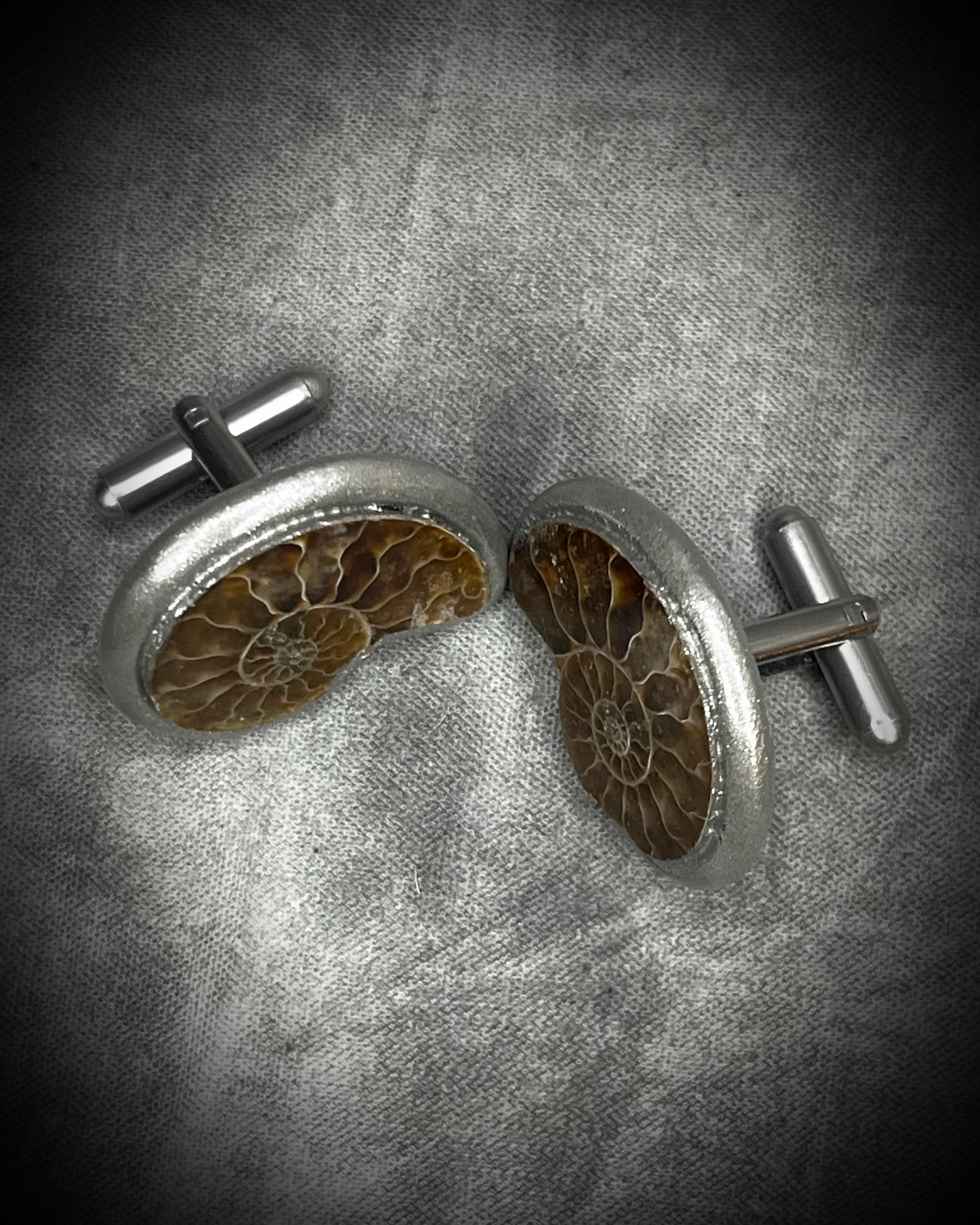 A pair of round cufflinks featuring detailed brown and gold ammonite fossils set in silver-tone metal, displayed on a textured gray background.