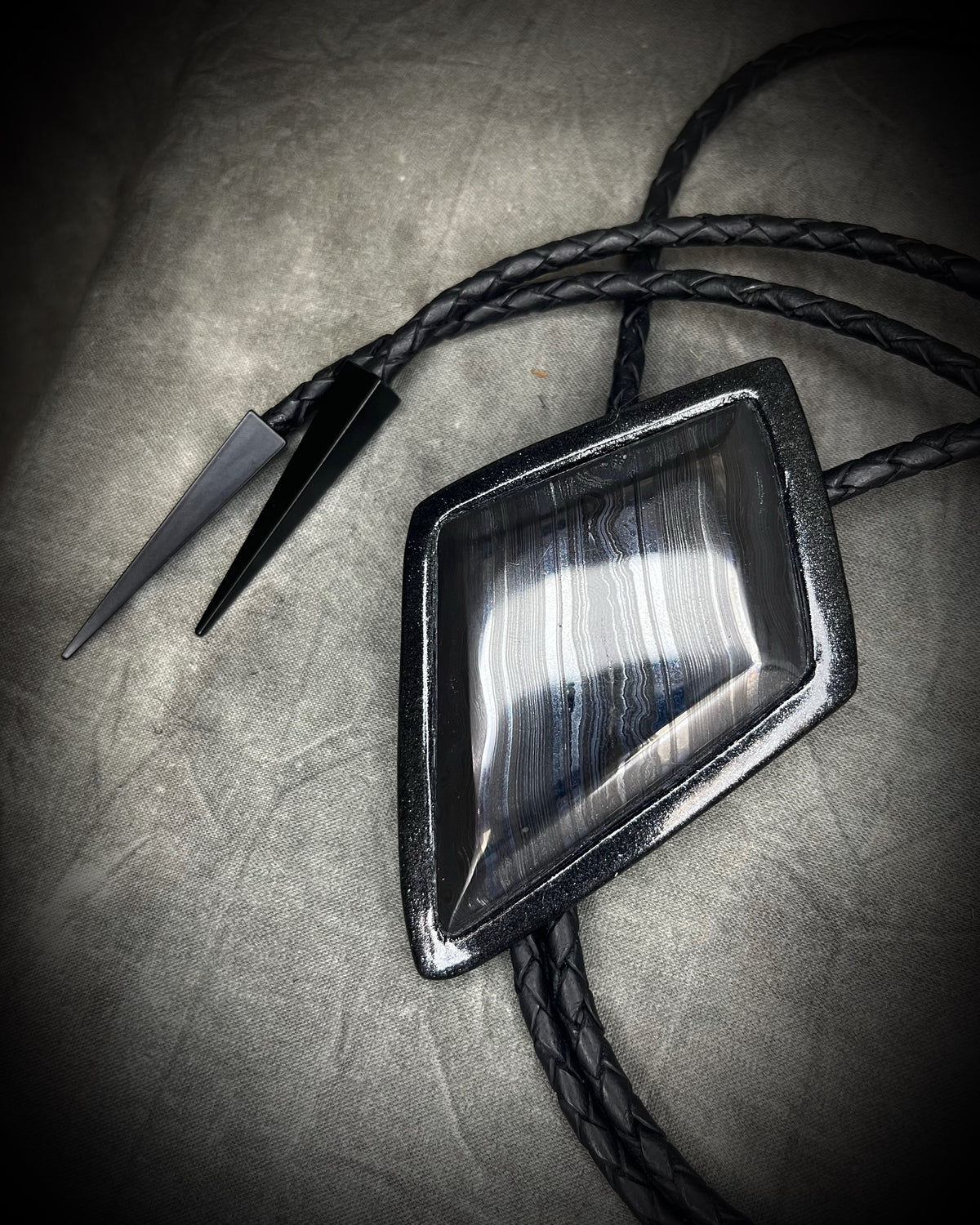 Genuine asymmetrical kite-shaped psilomelane bolo tie with black finish, black leather cord, and black spike bolo tips. Handmade by Become Spellbound.